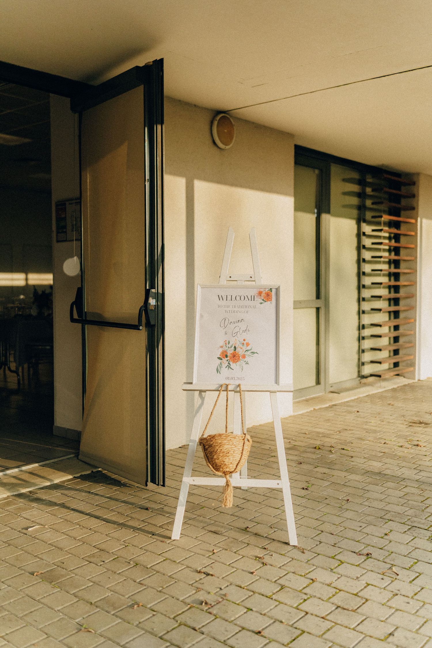 A welcoming white easel sign stand with a welcome poster displayed outside a building entrance in warm sunlight.
