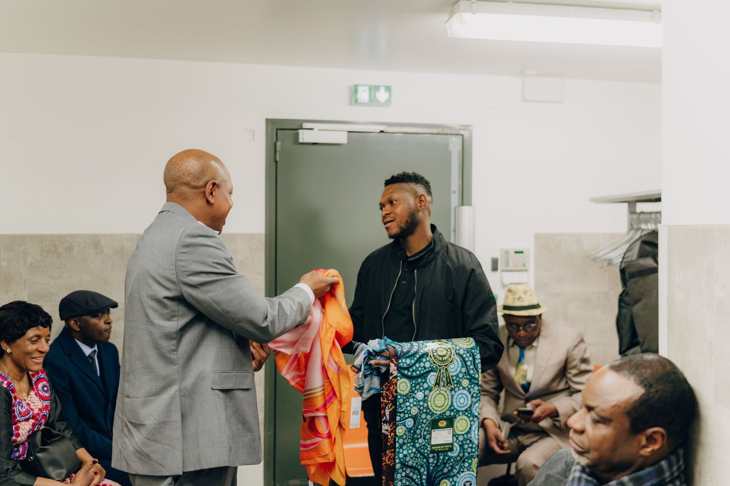 People interact in a meeting room with someone wearing an orange safety vest and colorful patterned shirt.