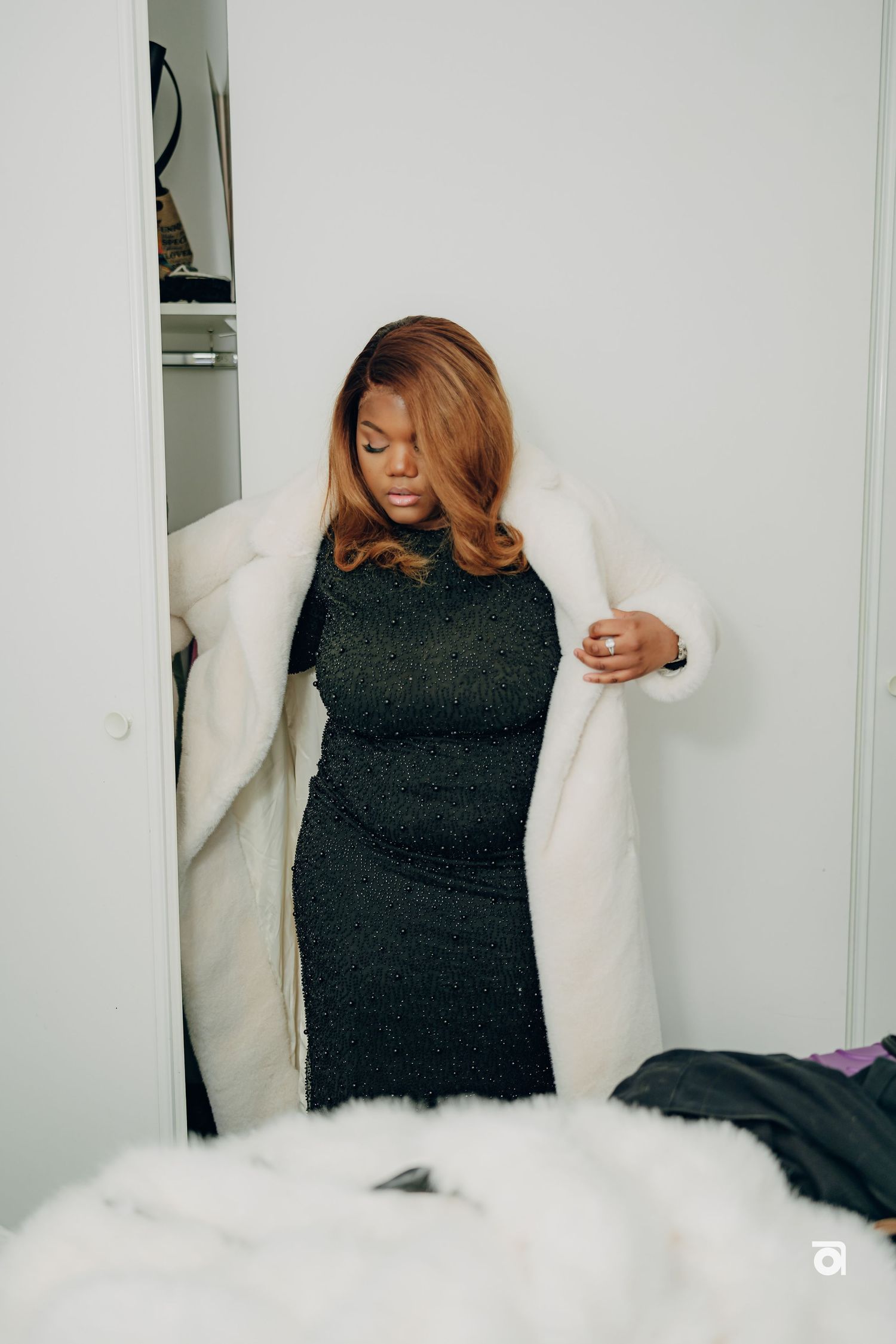 Woman in a black dress trying on a white winter coat in a minimalist room setting.