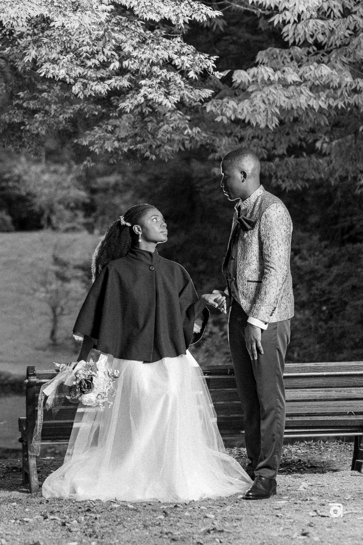 A black and white photograph shows two people in formal attire sharing a tender moment by a park bench under leafy trees.