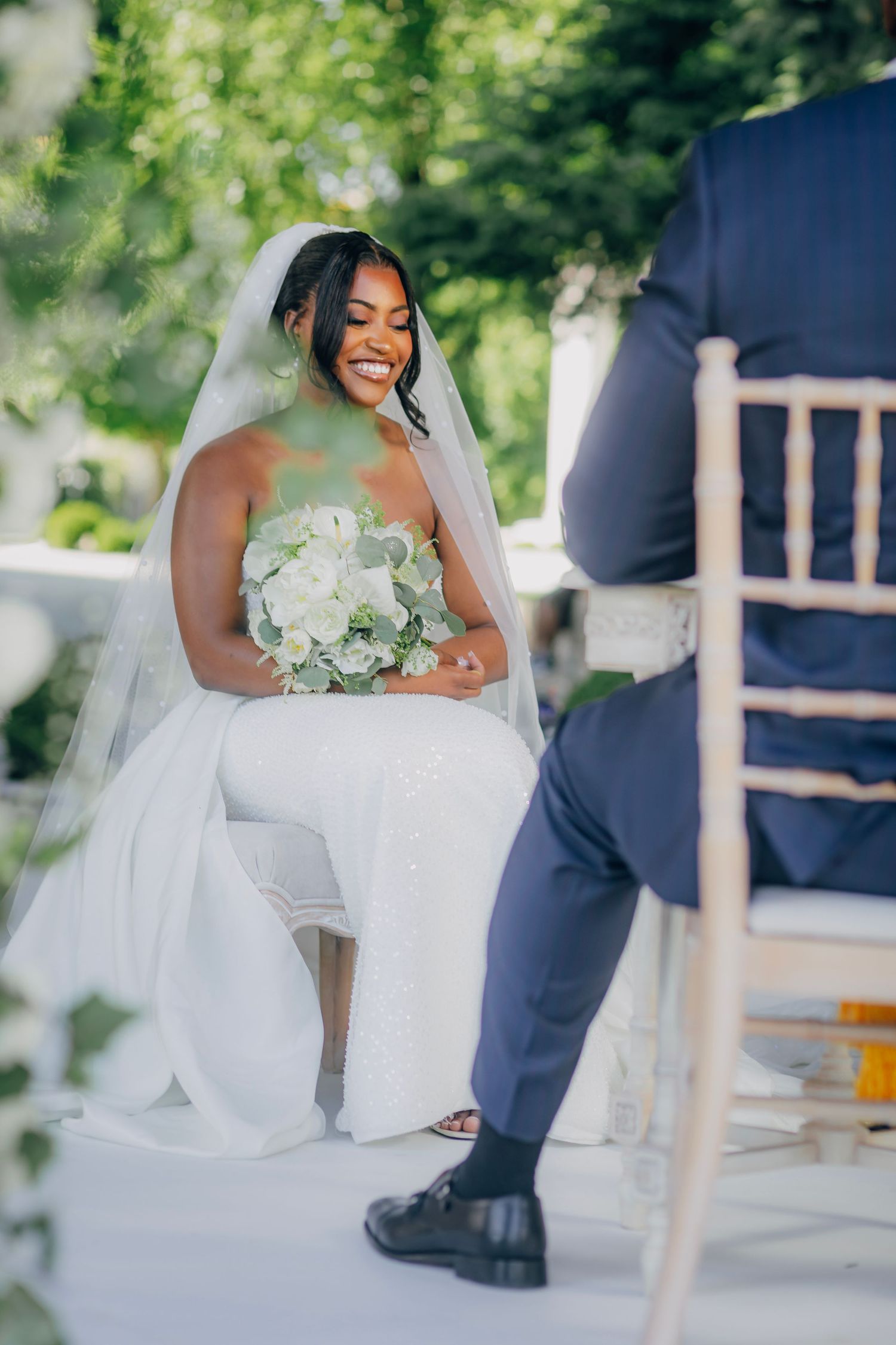 A bride in a stunning white gown holds a bouquet of white flowers during an outdoor ceremony.