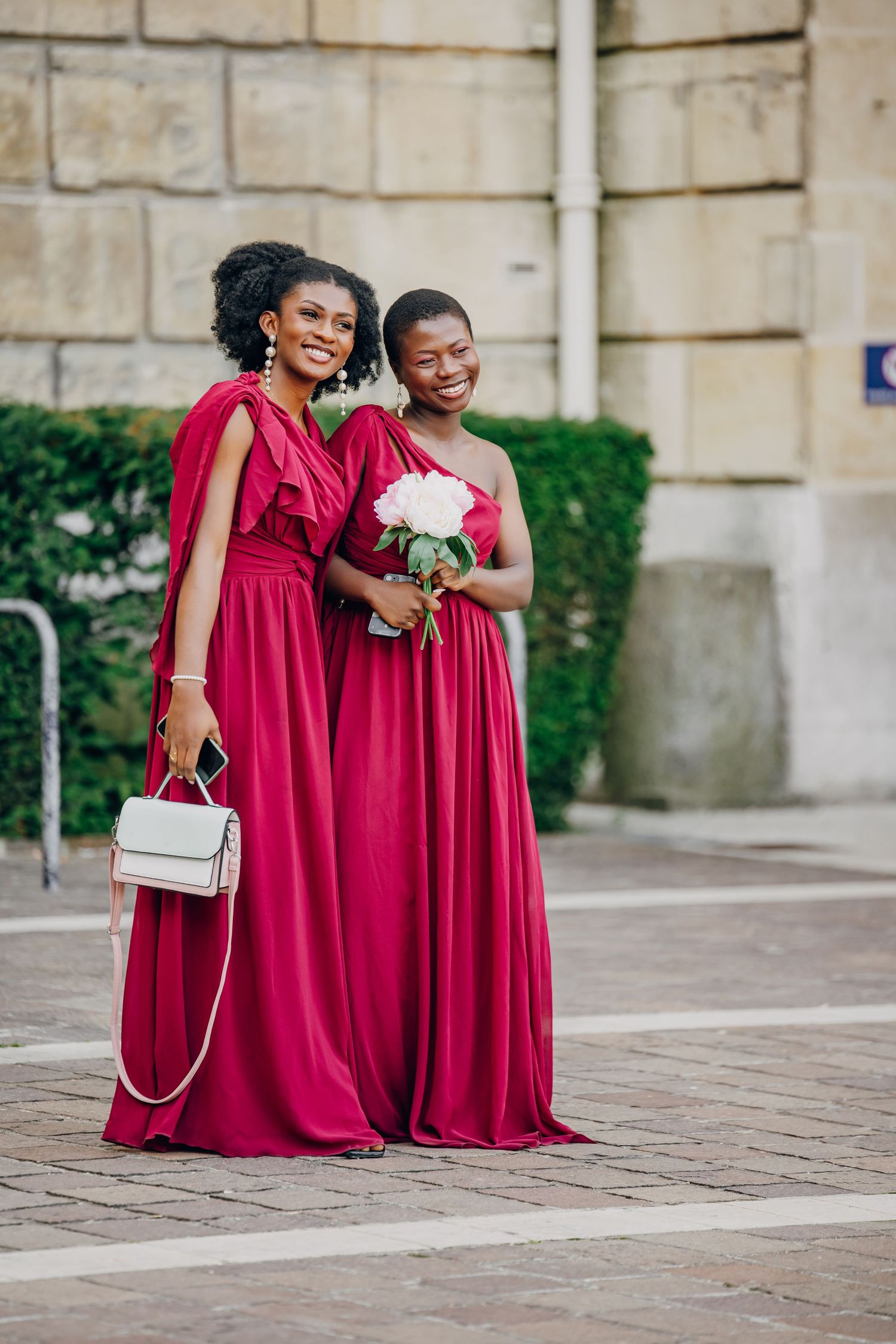 Two bridesmaids in matching floor-length red gowns pose together, one holding a white bouquet and one carrying a purse.