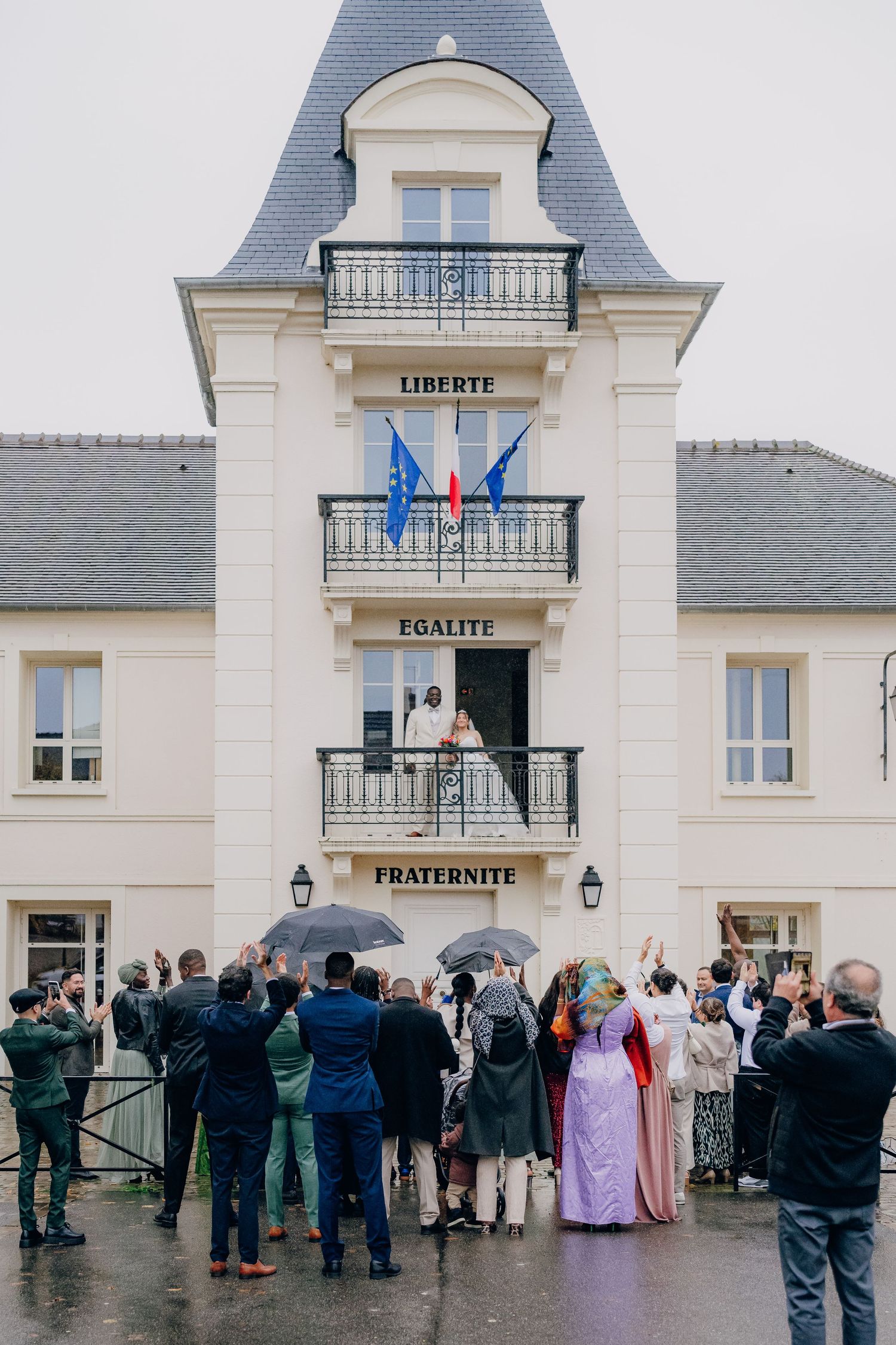 A crowd gathers outside a French-style mansion with balconies displaying Liberty, Equality, Fraternity signs and blue flags.