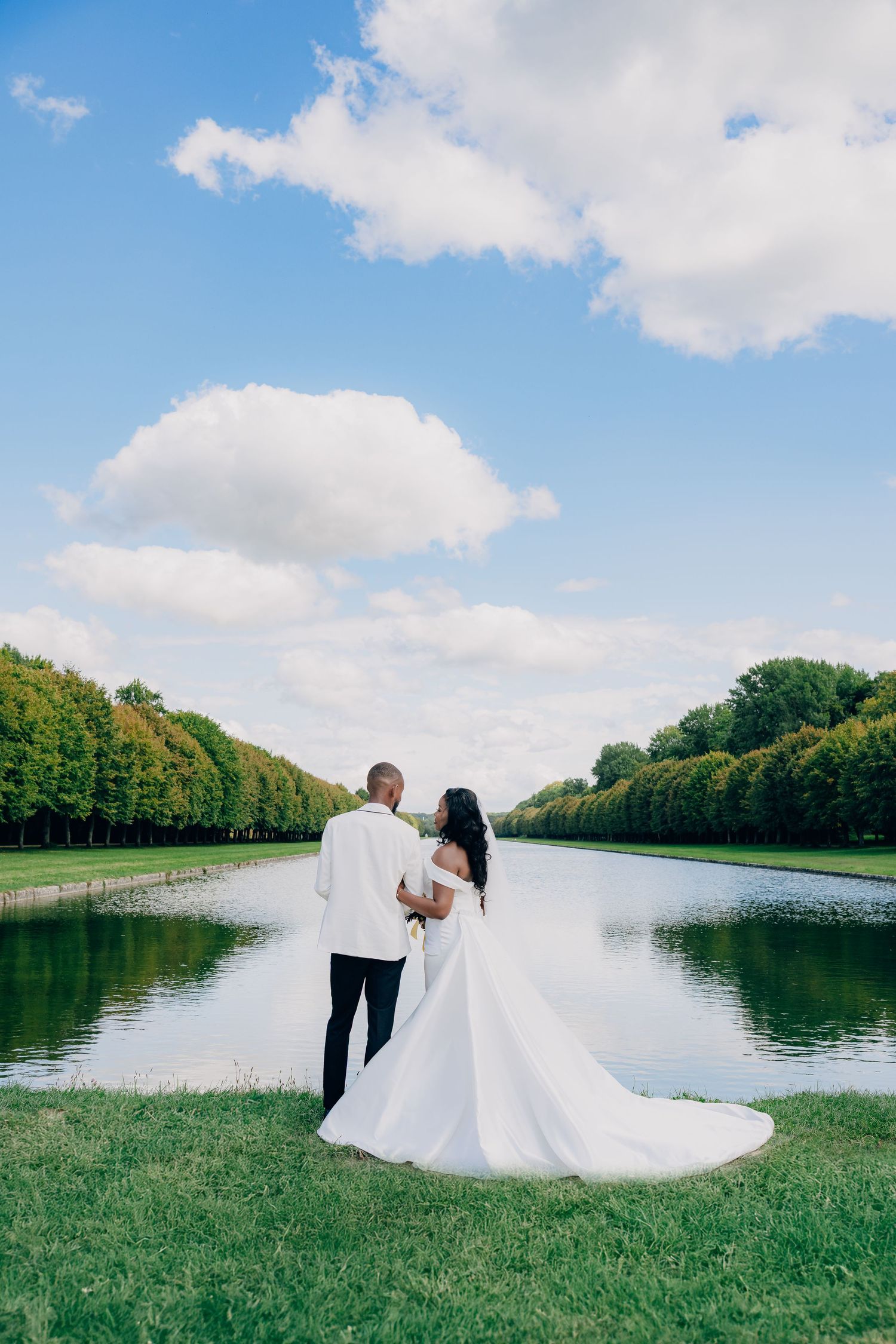 A couple in wedding attire stands by a serene reflecting pool surrounded by manicured hedges on a beautiful sunny day.