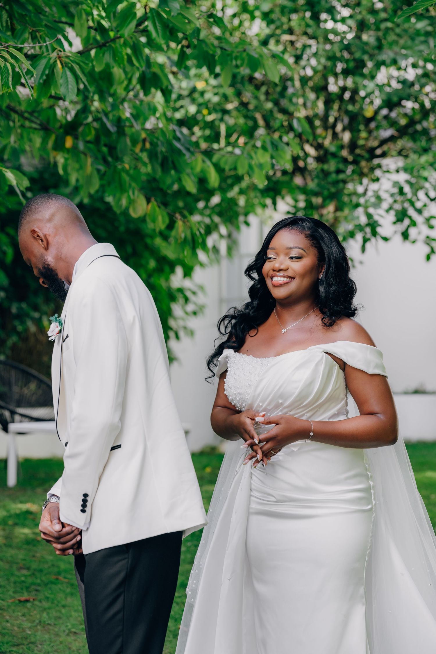 Wedding couple shares an intimate moment outdoors under green trees wearing elegant white attire.