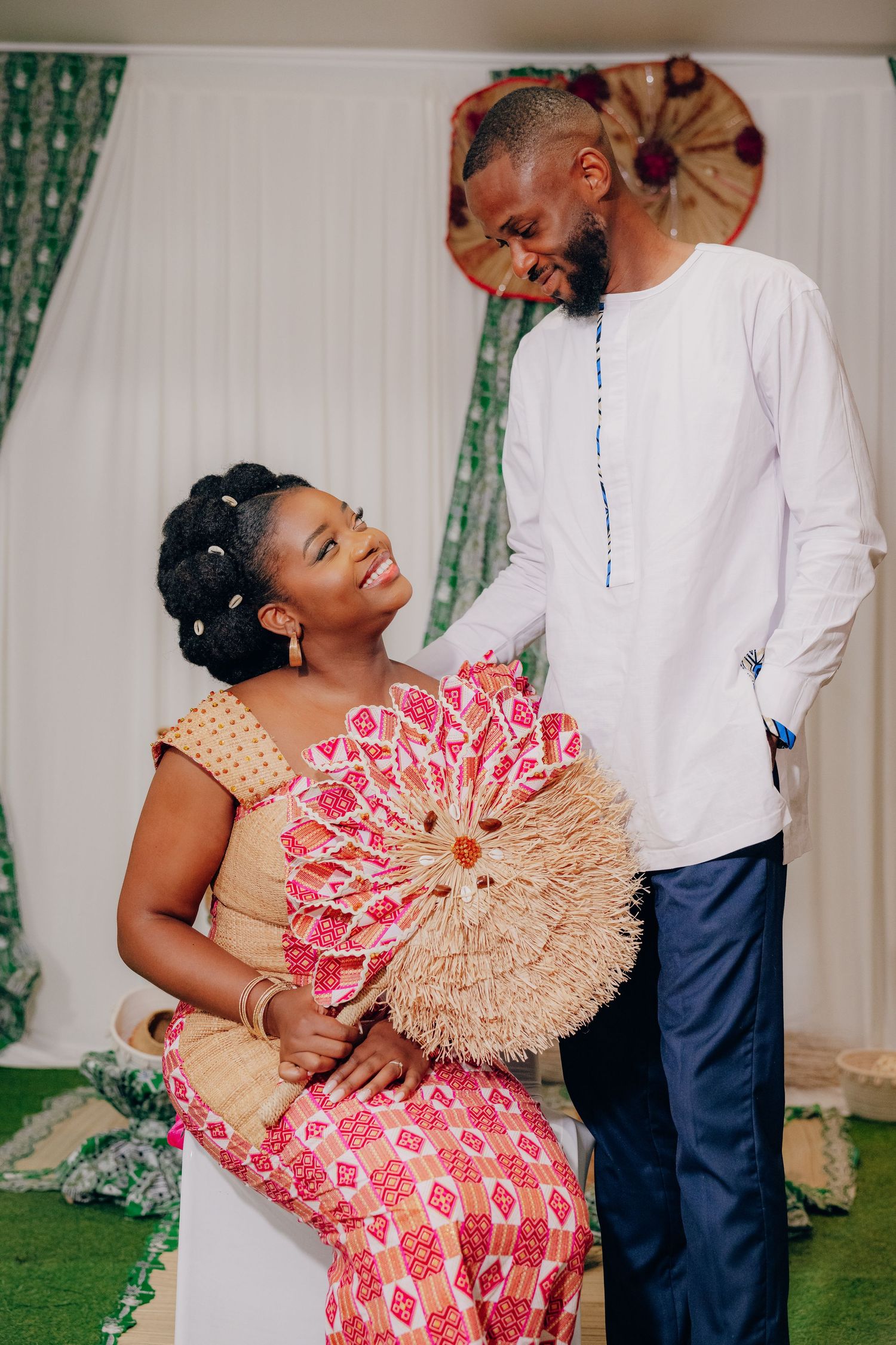 A couple shares a joyful moment with decorative fan and traditional attire at an indoor celebration.