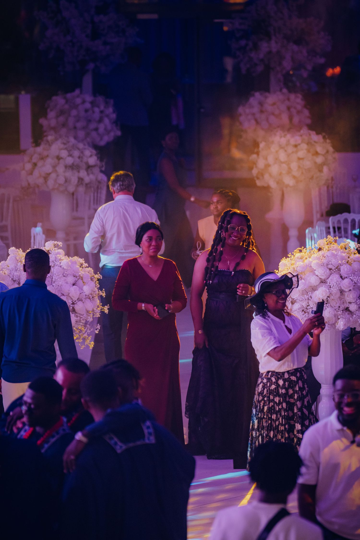 Wedding reception guests mingle and dance in a dimly lit ballroom decorated with white floral arrangements.