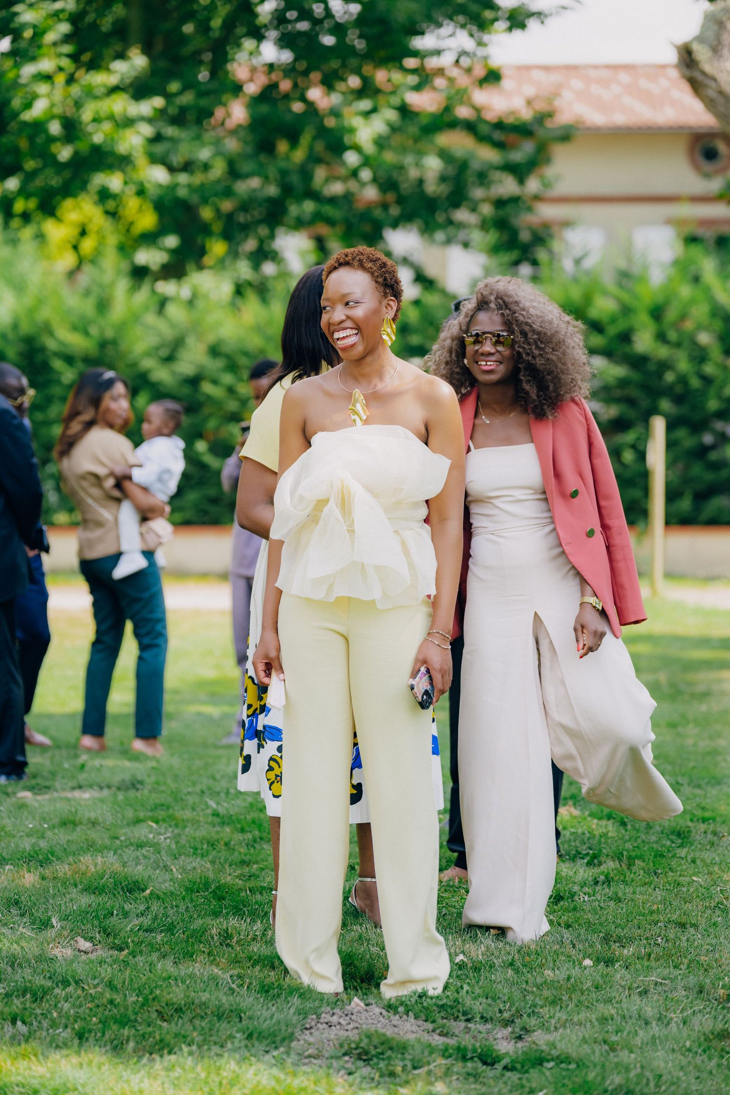 Stylish wedding party members in light colored outfits laugh together on grassy grounds.