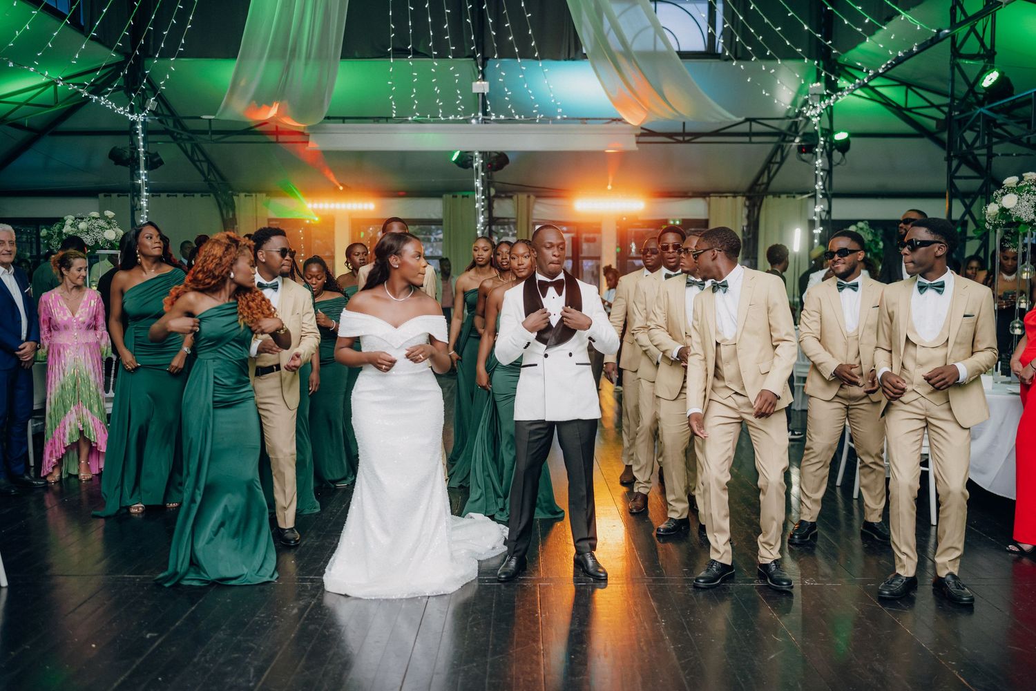 Wedding party lined up on dance floor with bridesmaids in green dresses and groomsmen in beige suits.
