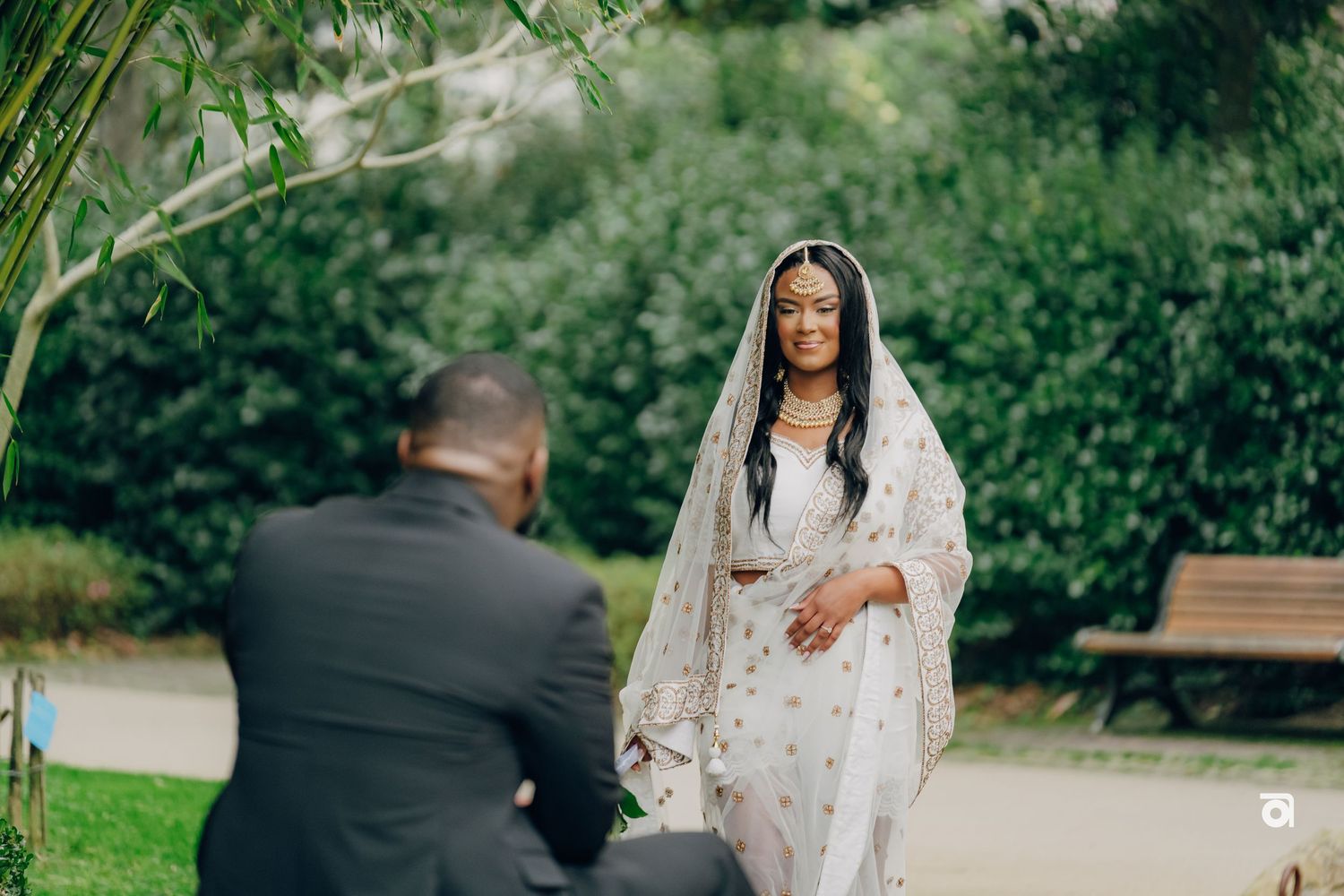 A romantic proposal moment captured in a lush garden setting with greenery and a wooden bench in the background.