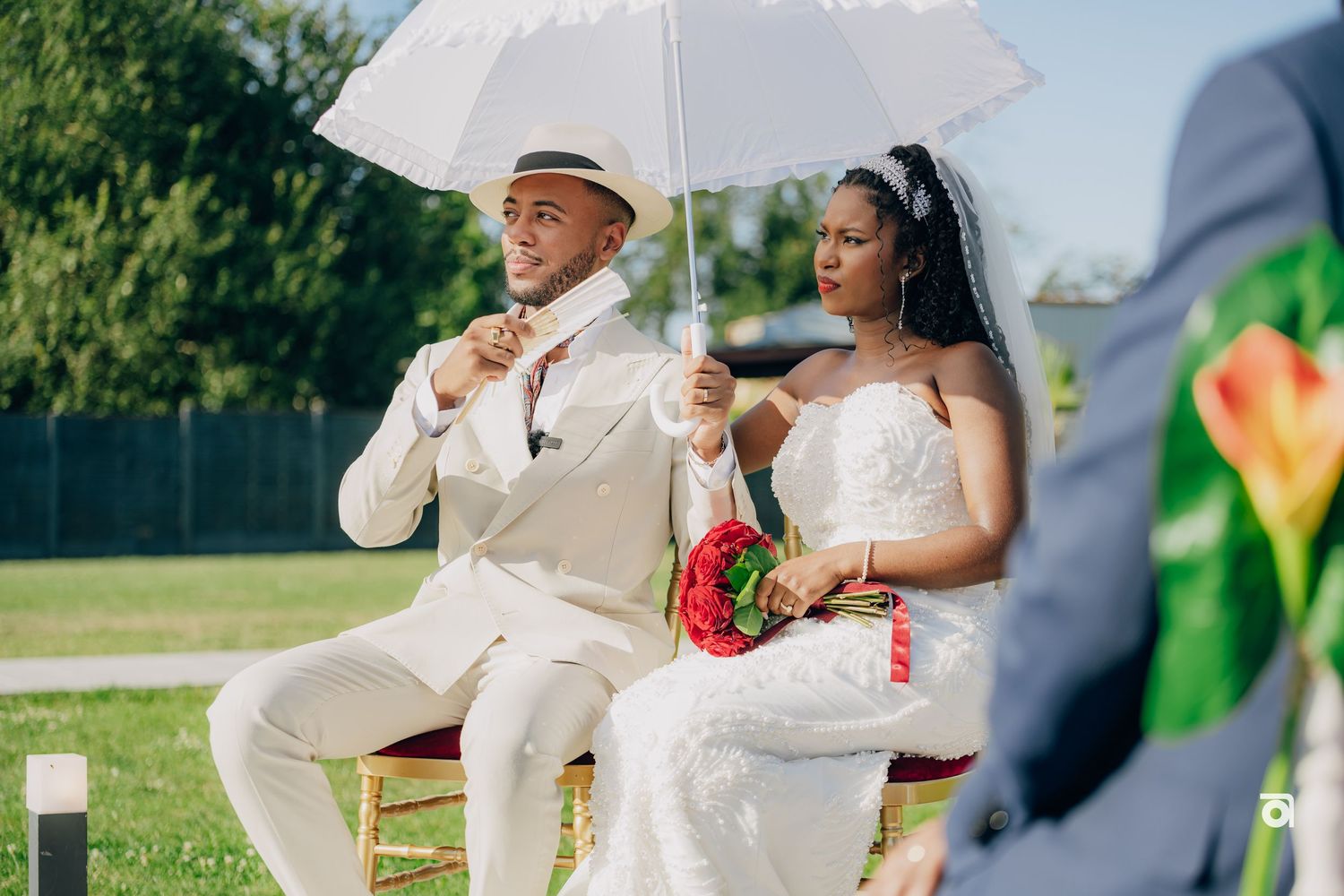 A couple sits under a white umbrella during an outdoor wedding celebration on a sunny day.