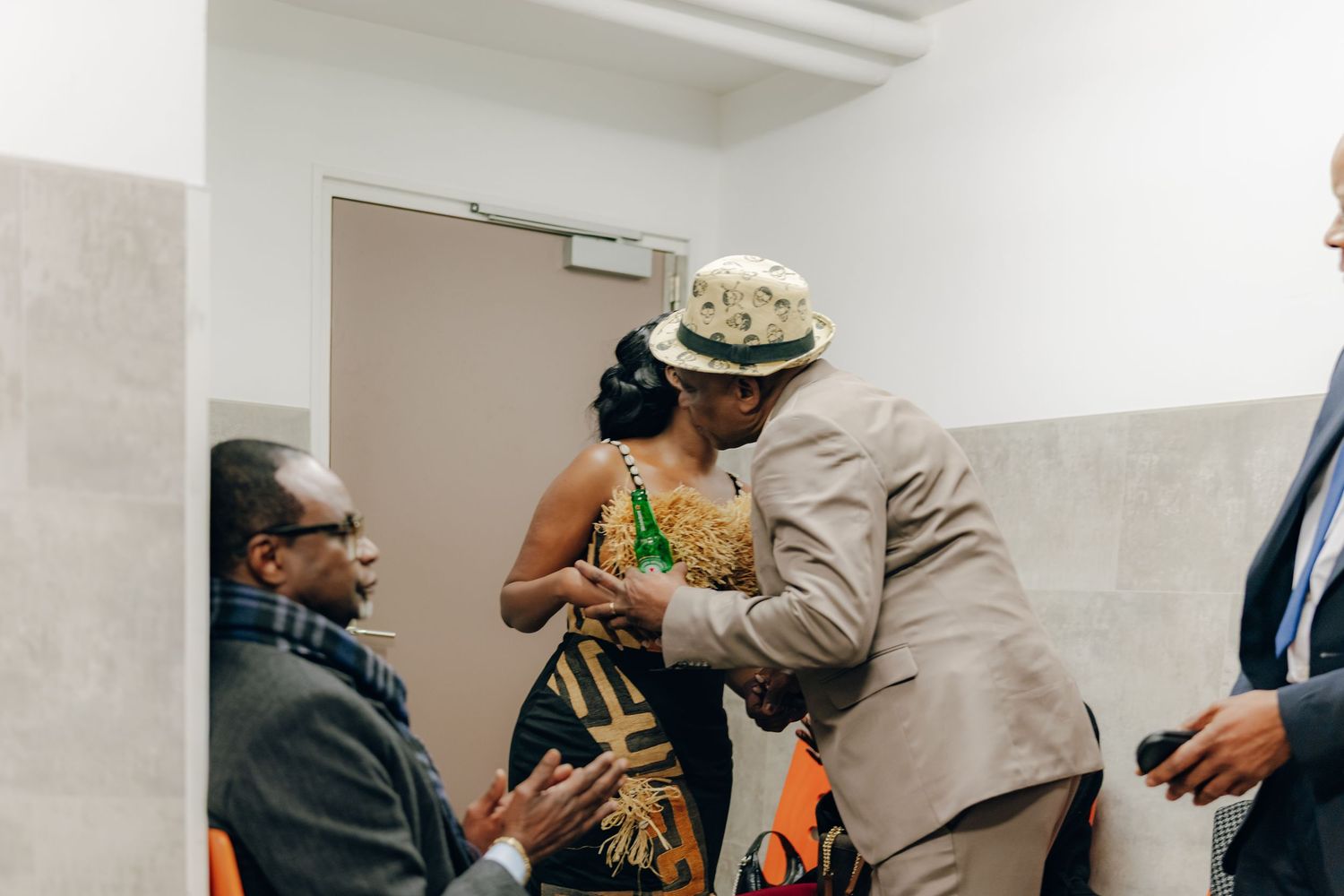People interacting in a hallway with one in formal attire wearing a beige suit and hat.