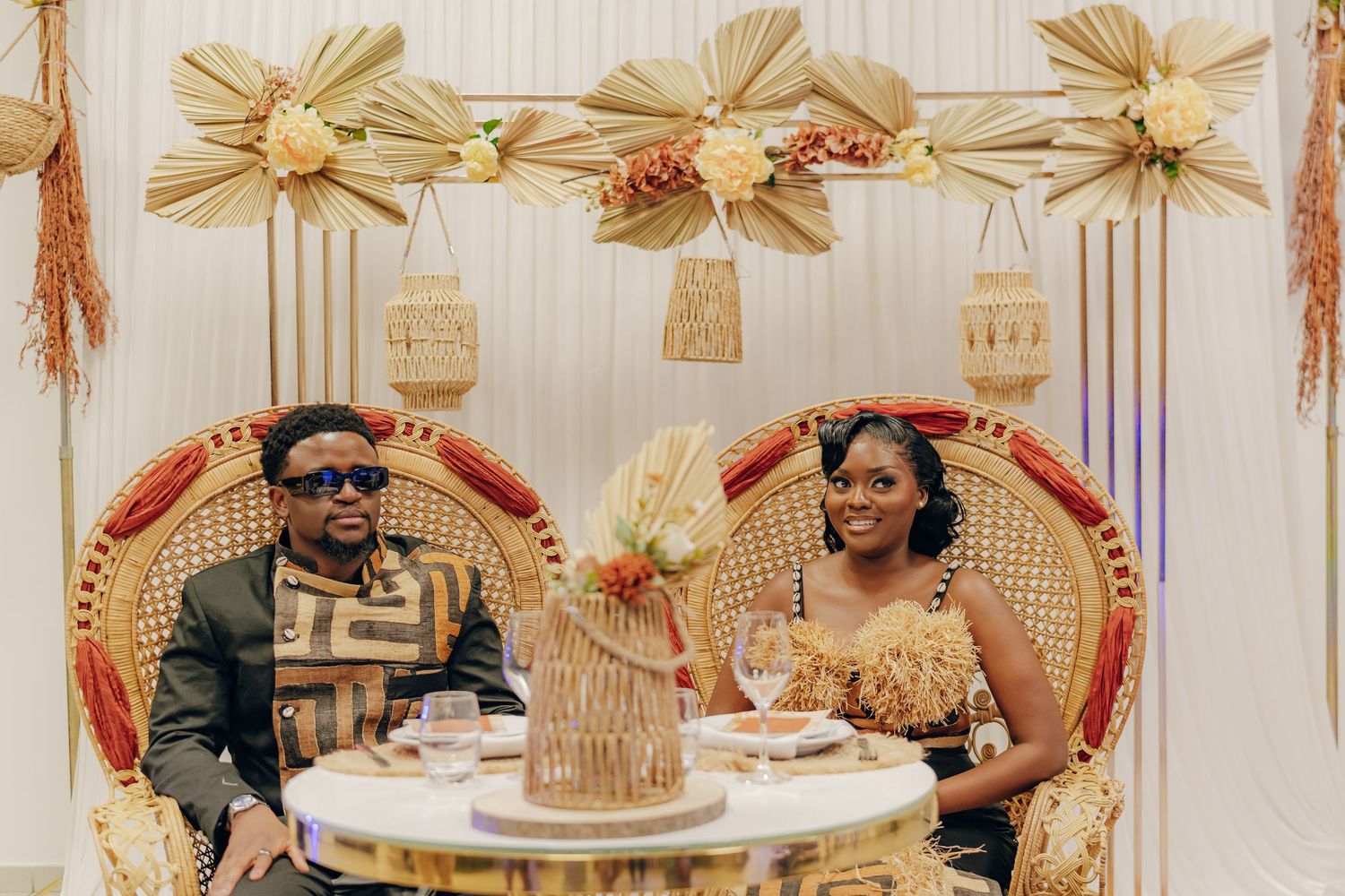A couple enjoys cake and drinks at their elegant peacock chair wedding setup with boho style decor and floral arrangements.