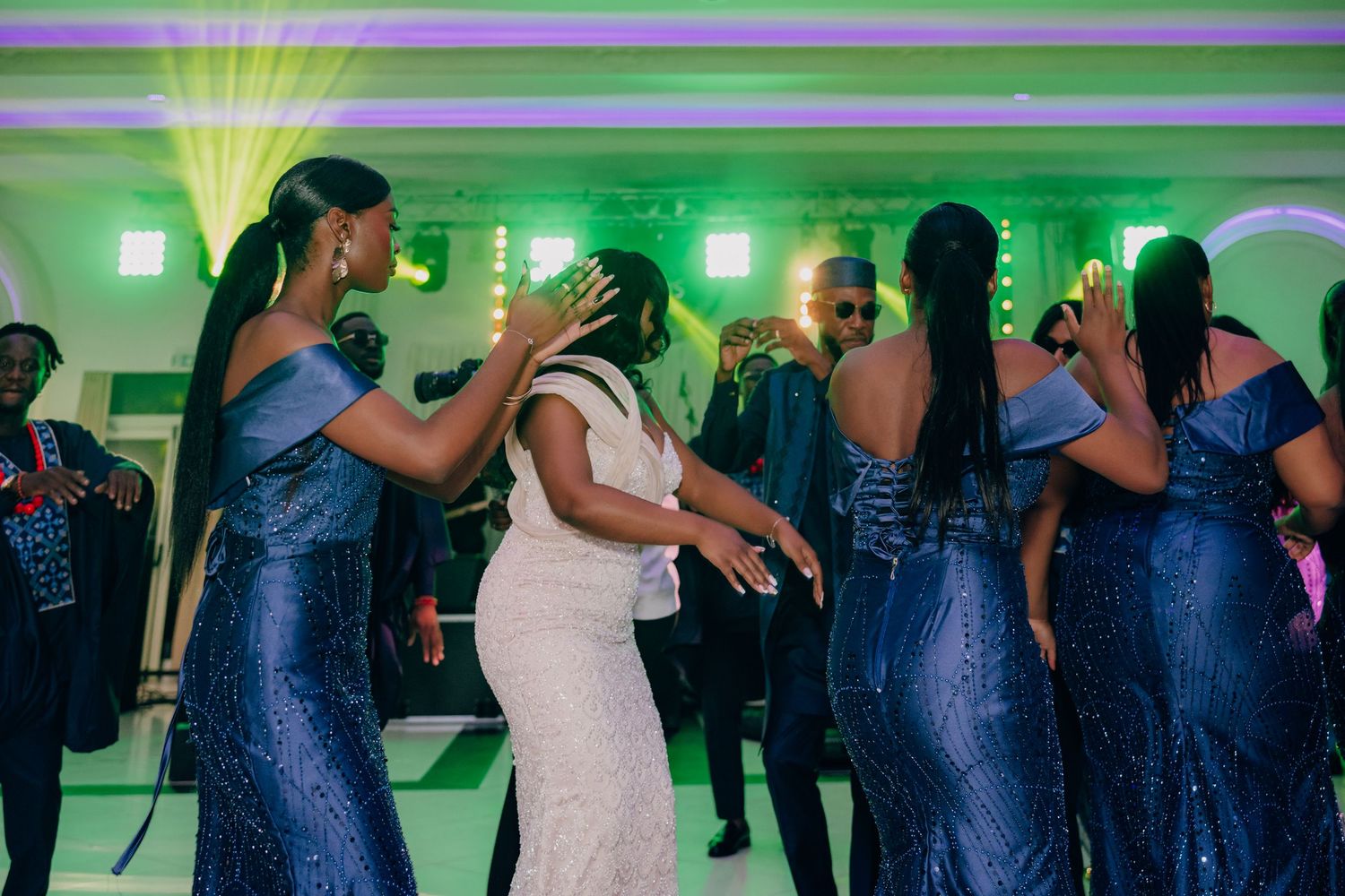 Wedding celebration with guests dancing in blue dresses under green lighting at a reception venue.