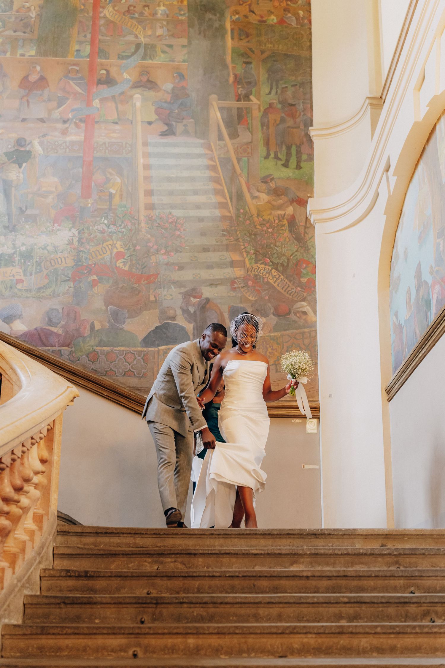 A couple ascends an elegant staircase with ornate wall murals in a historic building.