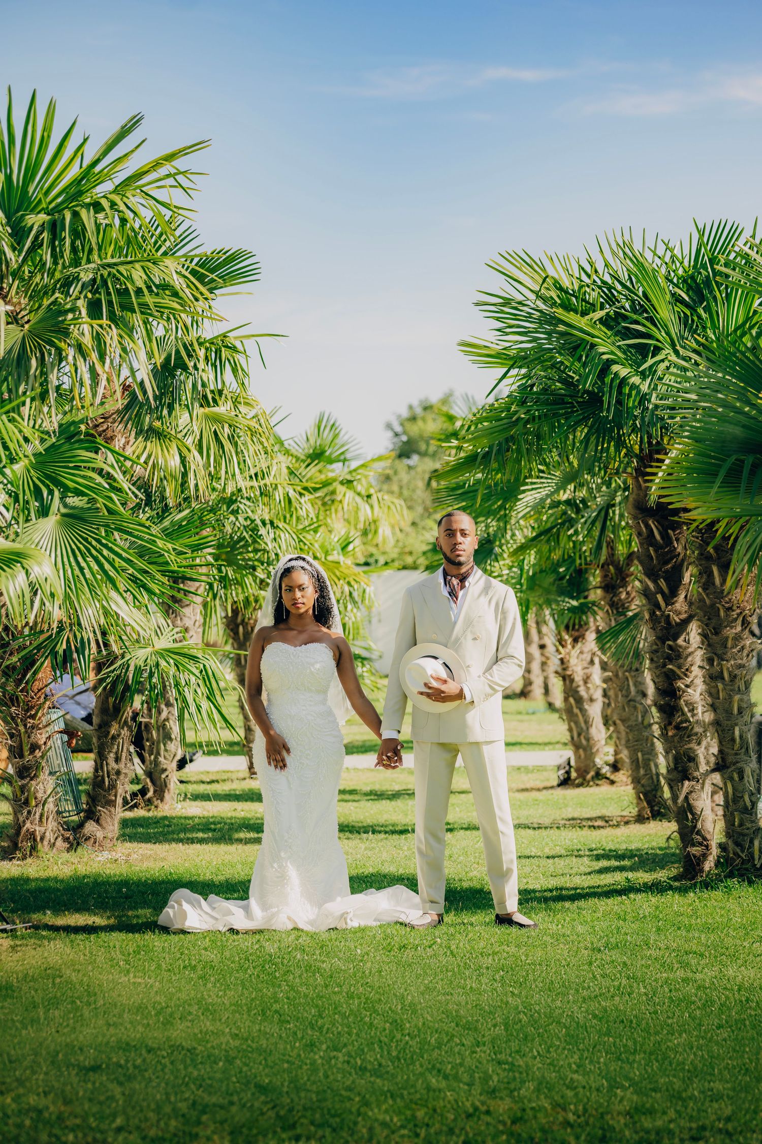 A couple in wedding attire stands among palm trees on a manicured lawn under blue skies.