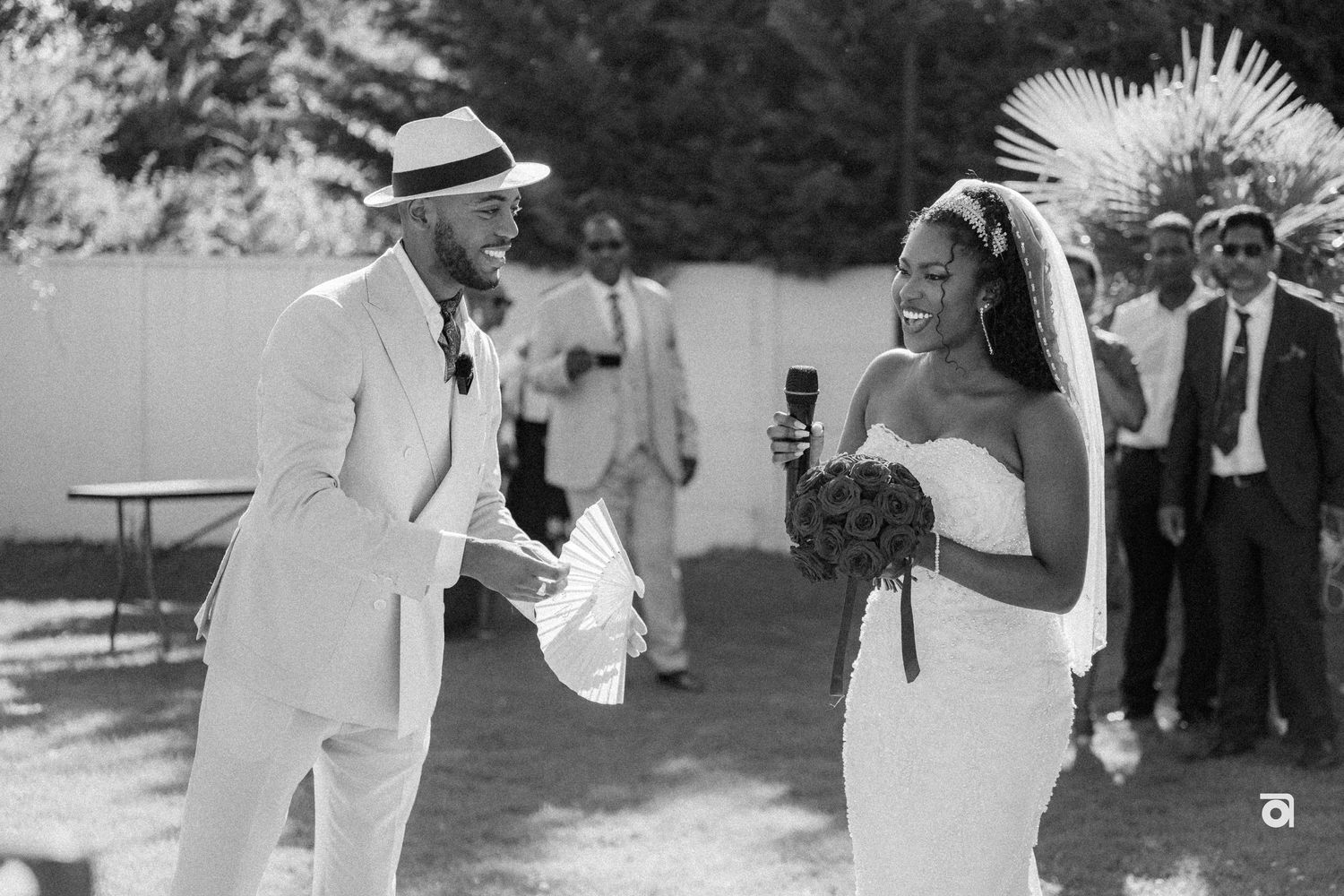 A black and white photo of a wedding celebration with guests in formal attire exchanging smiles outdoors.