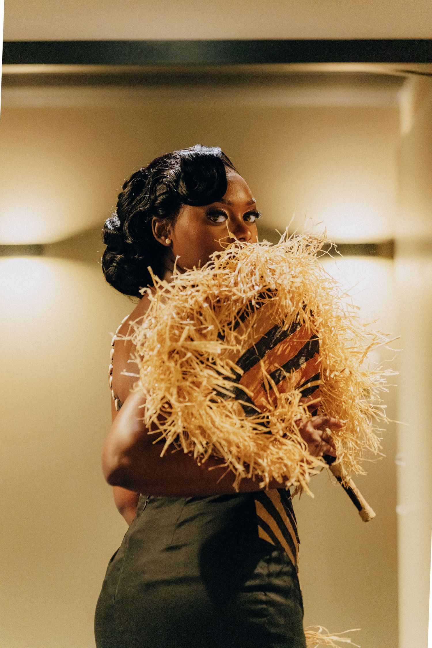 A person in elegant attire holds a golden straw fan while posing against a neutral background.