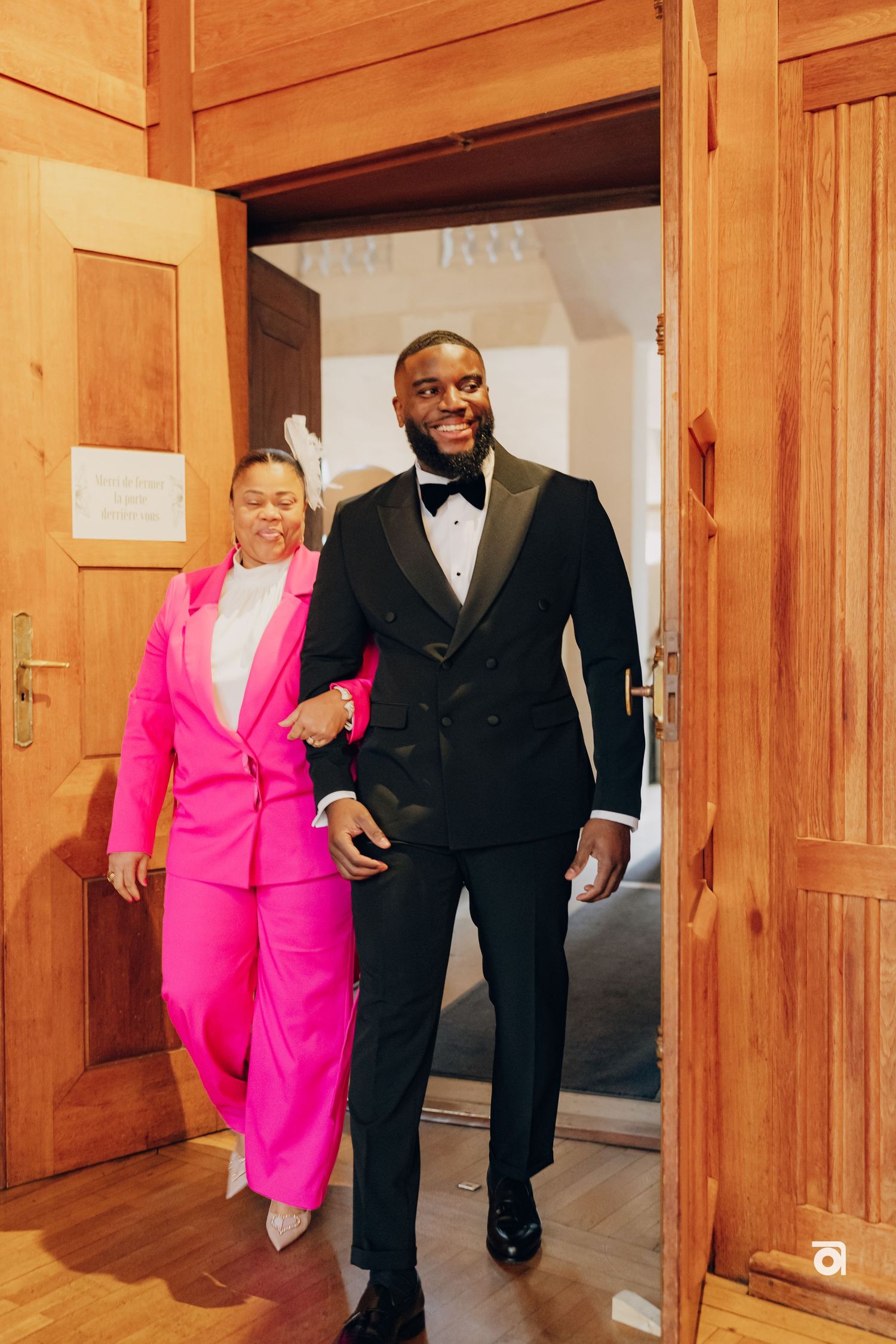 A couple wearing formal attire walk through a wooden doorway, with one in pink and one in a black tuxedo.