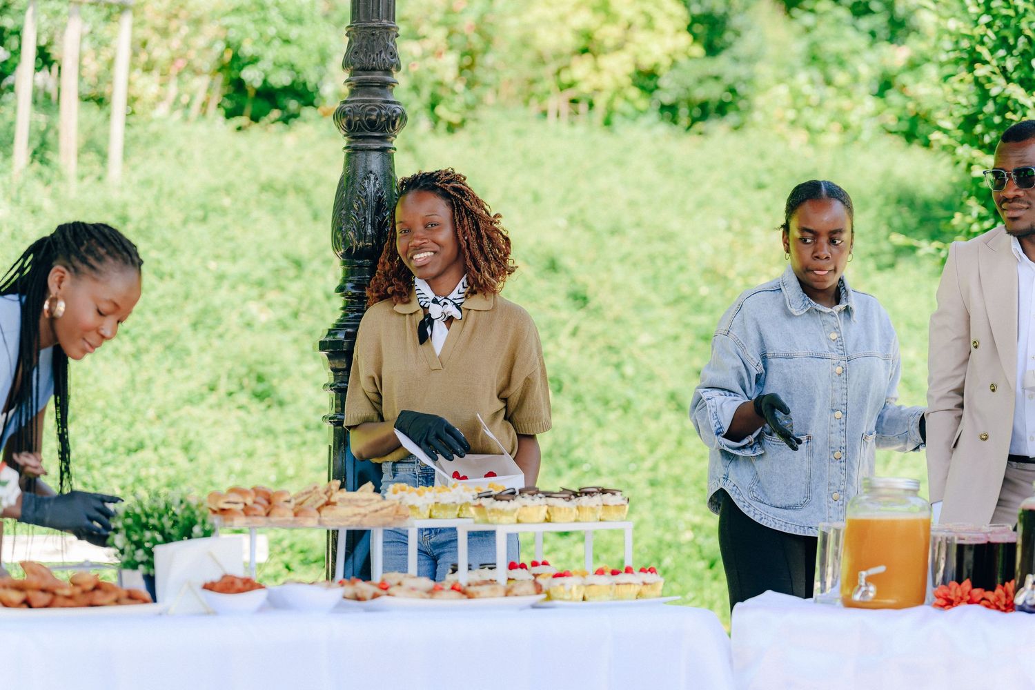 People gather around an outdoor buffet table with food and drinks during a catering event in a green garden setting.
