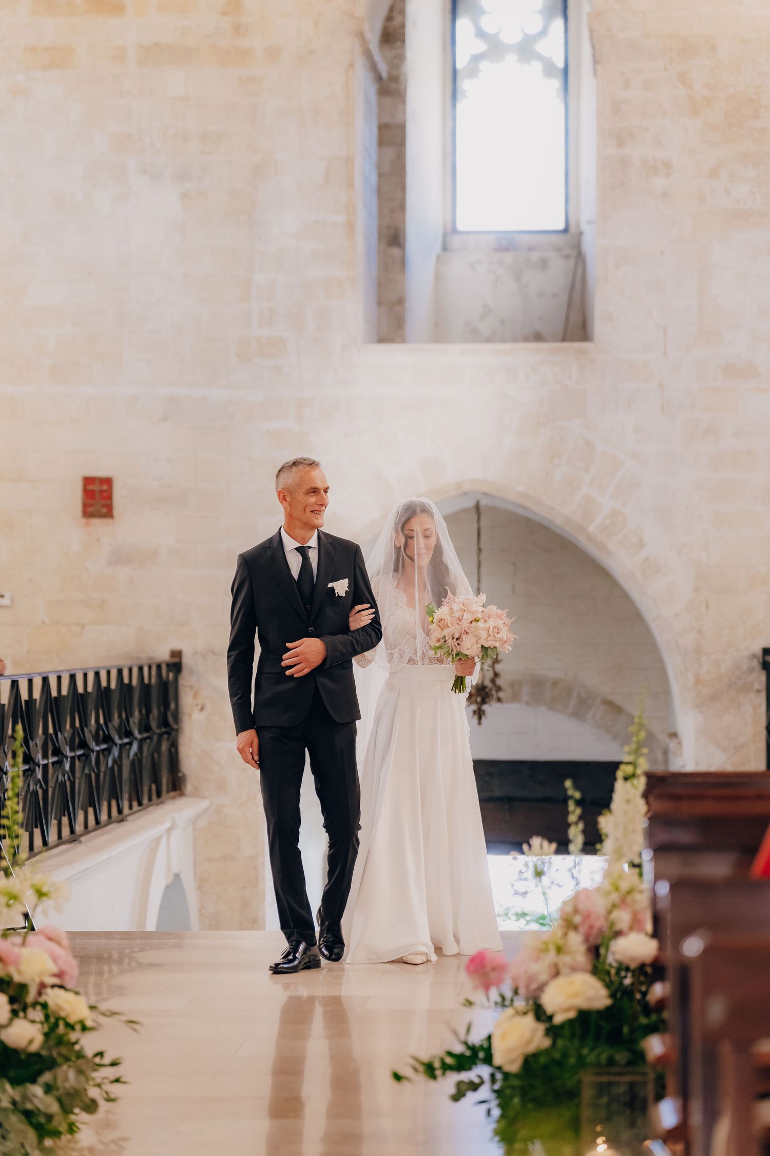 A wedding ceremony in a light-filled stone church with floral arrangements lining the aisle.