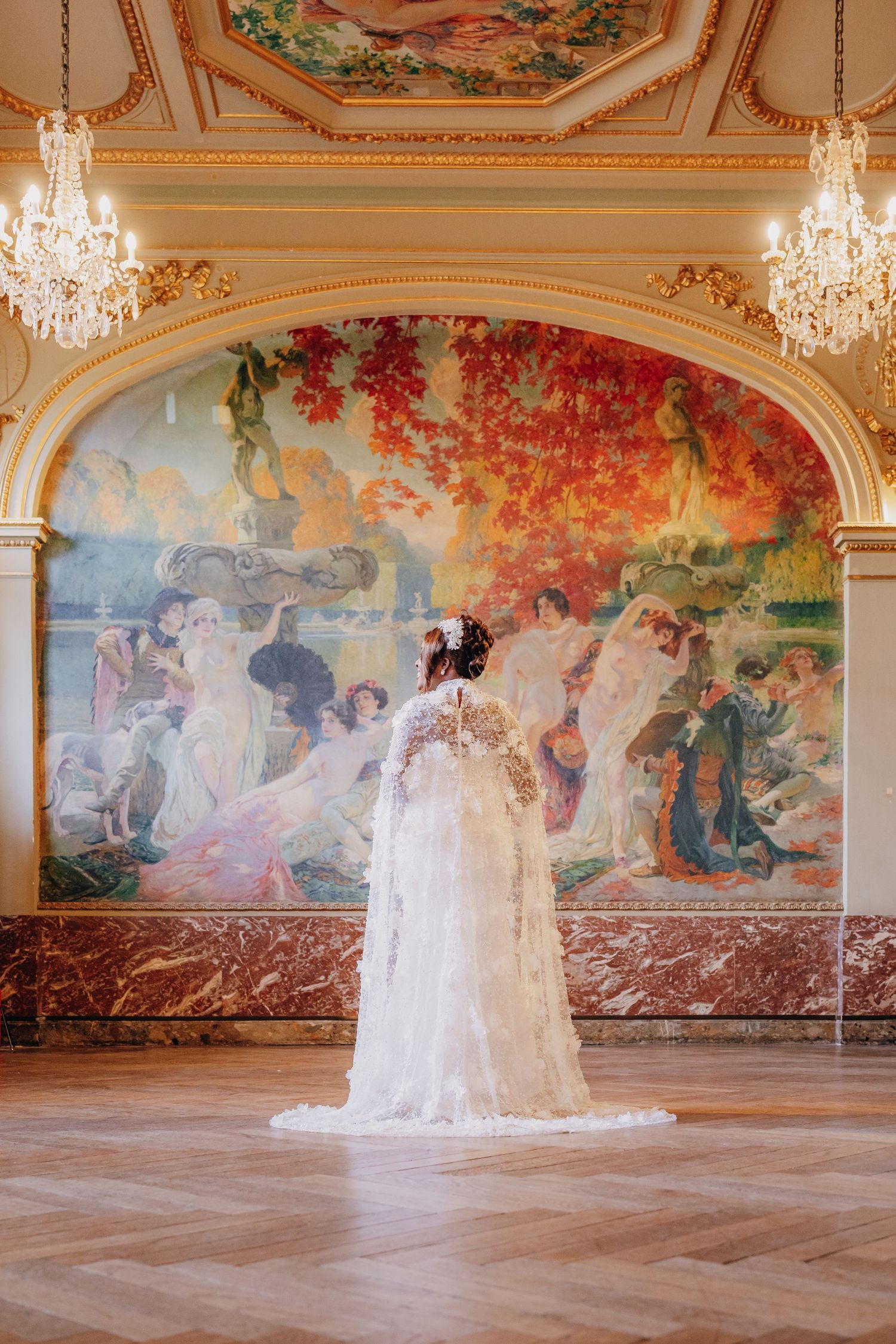 A figure in a flowing white wedding gown stands in an ornate ballroom with crystal chandeliers and painted wall murals.