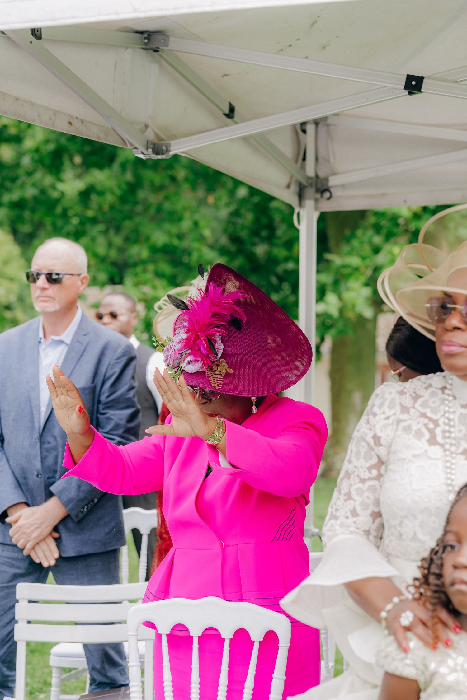 Wedding guests in bright attire including a vibrant pink hat seated under white tents.