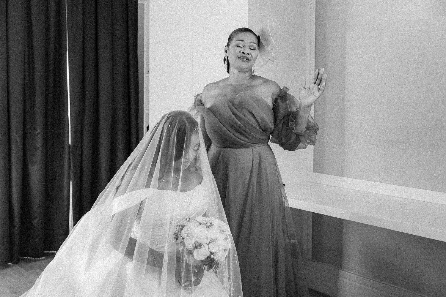 A person in an elegant white wedding gown and veil poses in a room with dark curtains.