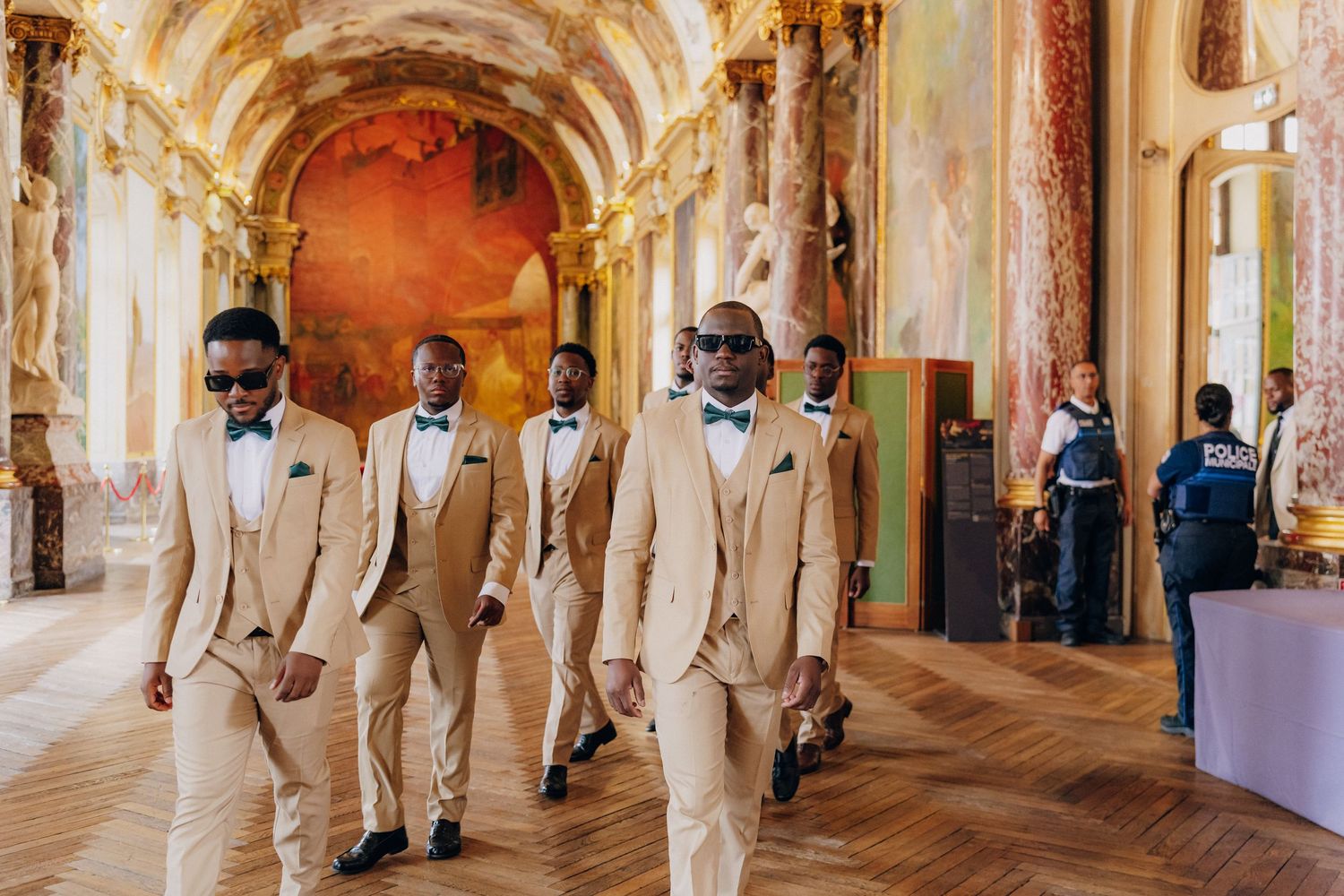 A wedding party in white suits walks through an ornate baroque hallway with gilded arches and frescoed ceilings.