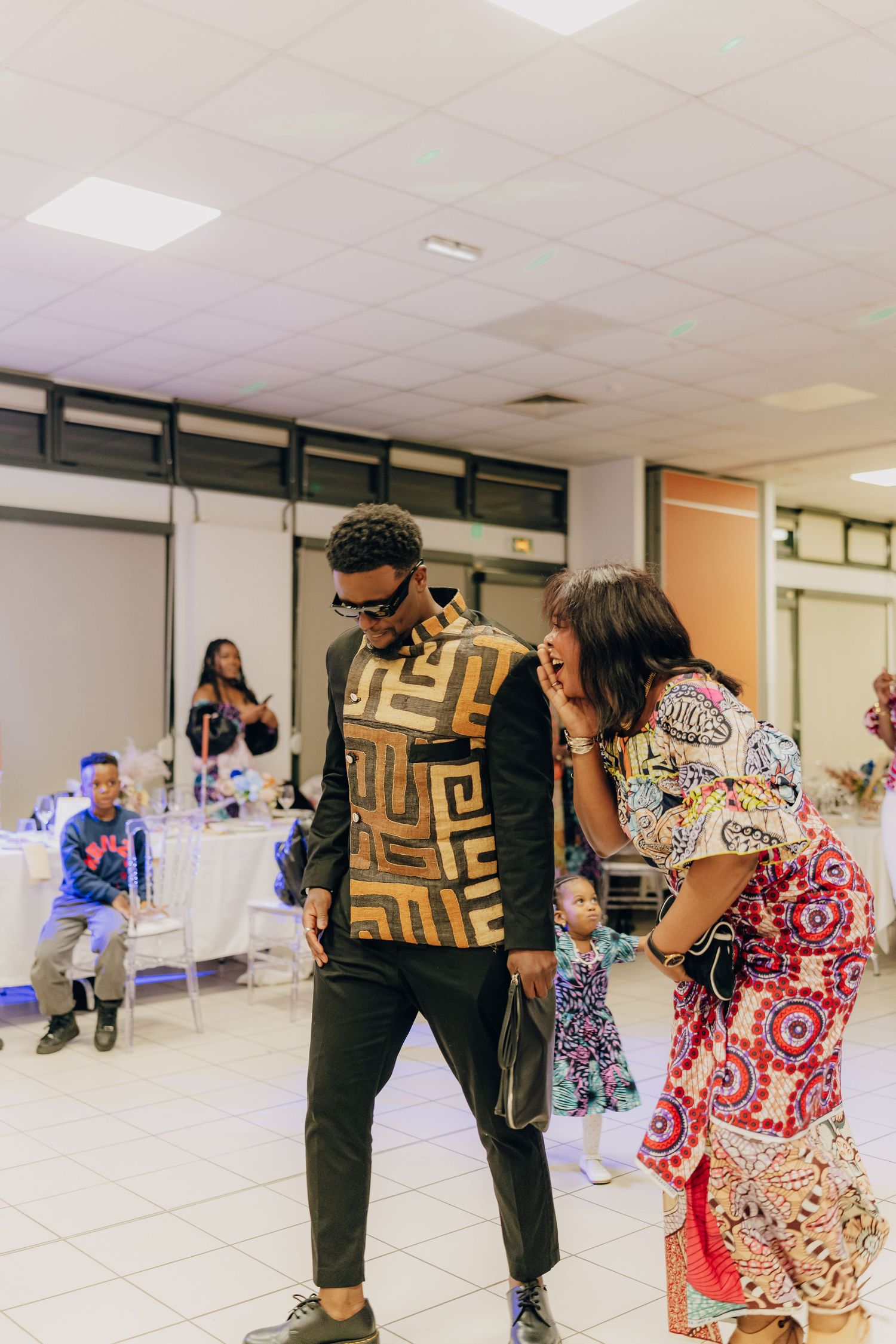 People dance and socialize at a party in a community hall with white fluorescent lighting.