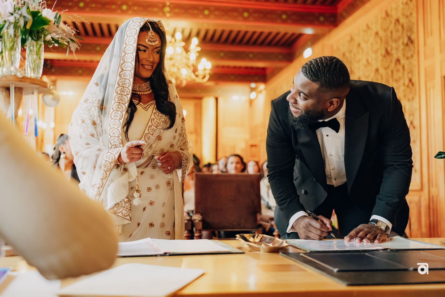 Couple signing wedding documents in an ornate room with gold wallpaper and chandelier lighting.