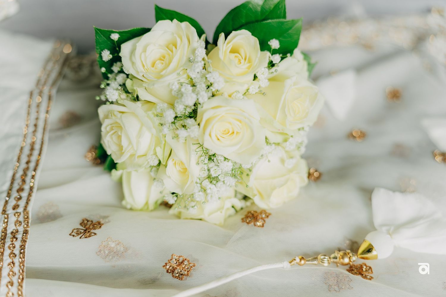 A delicate bridal bouquet of cream white roses and baby's breath rests on embroidered fabric with gold accents.