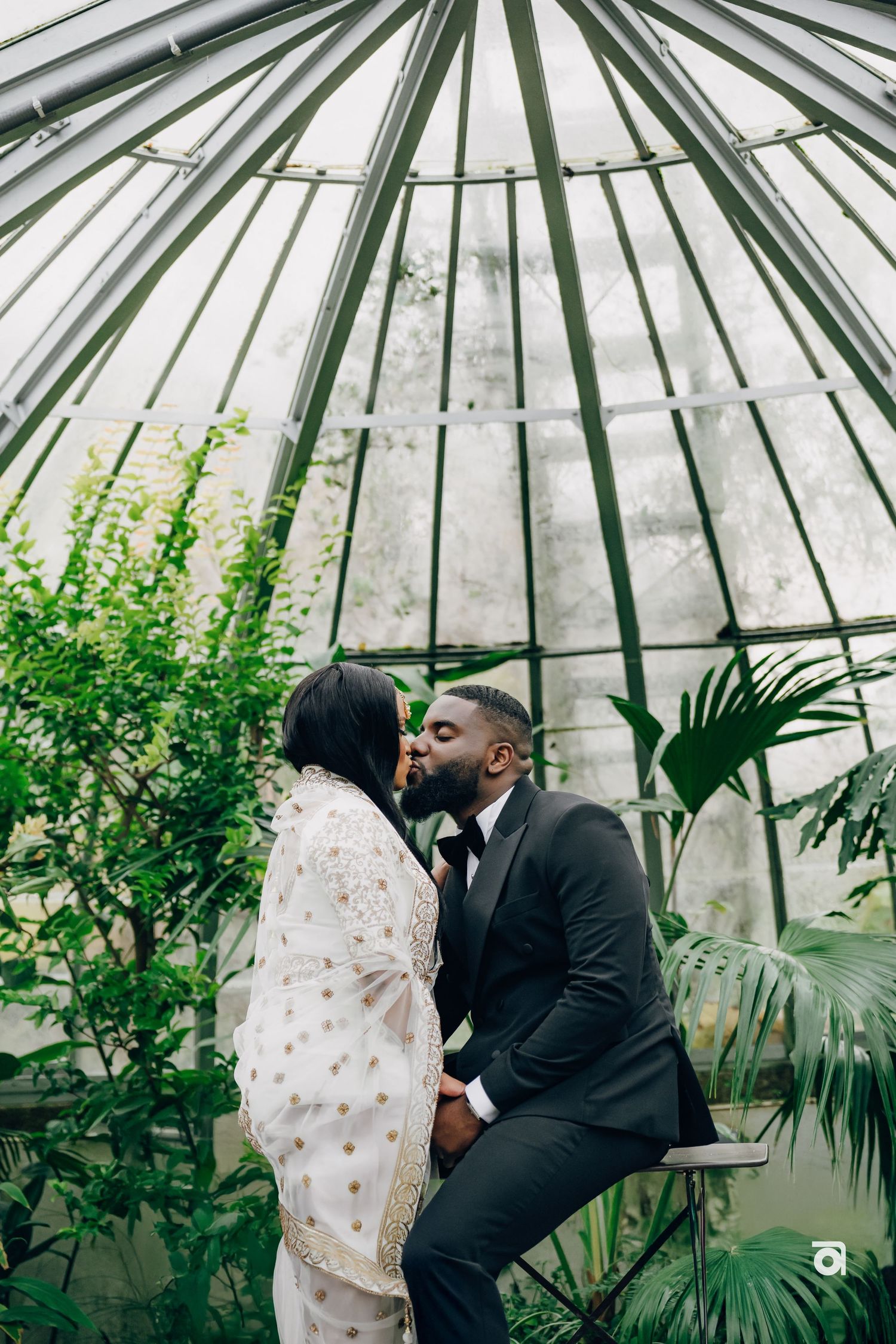 A romantic moment in a greenhouse with lush plants and glass ceiling architecture.