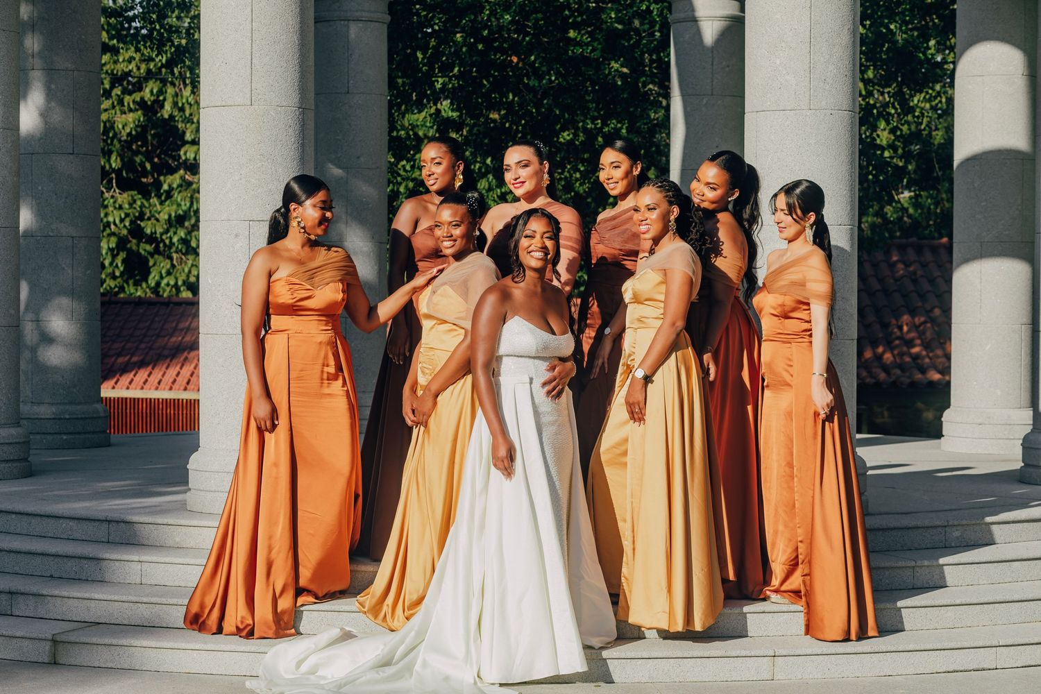 Bridesmaids in flowing orange and gold dresses pose with bride in white gown against classical columns.