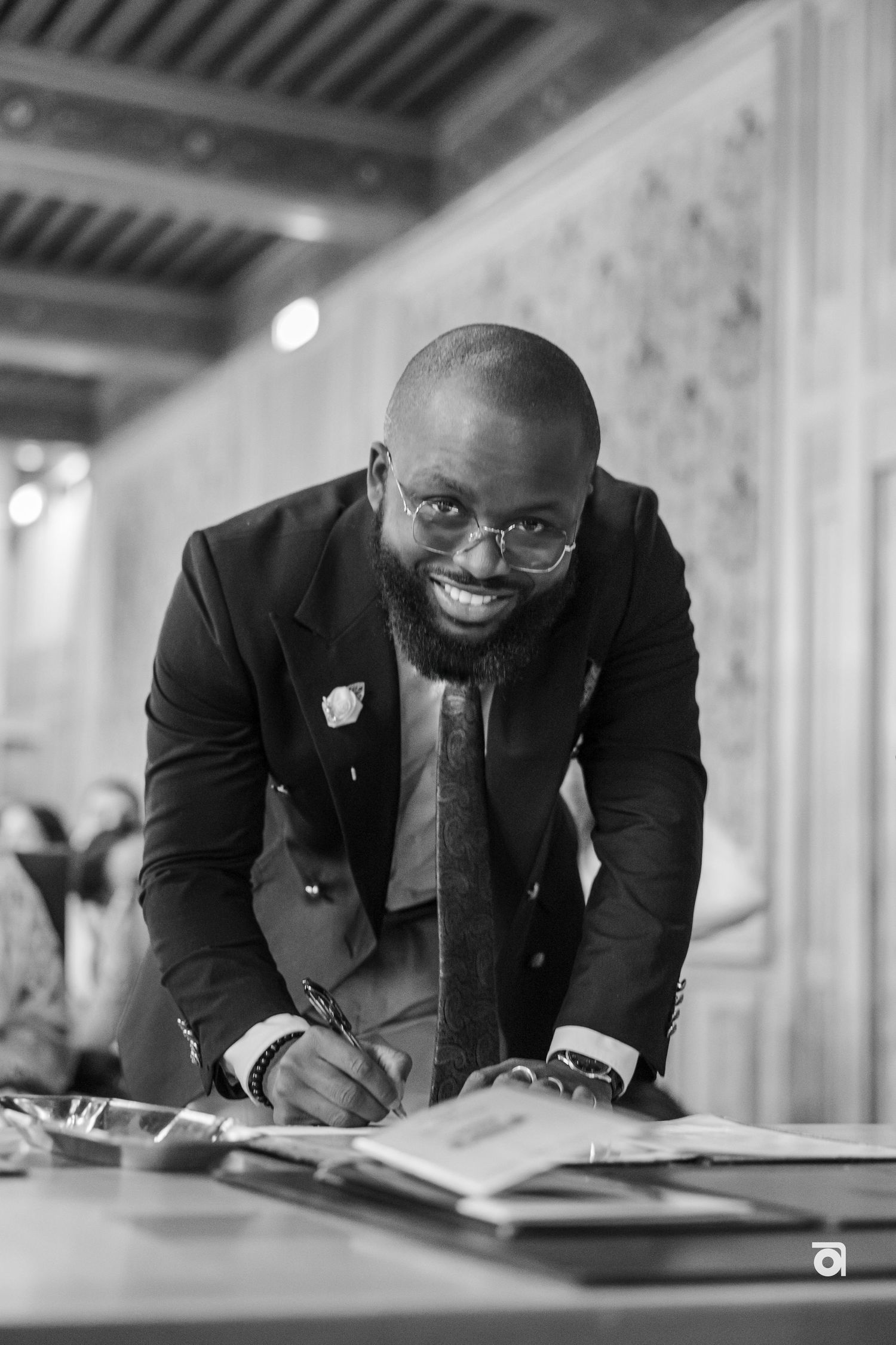 A smiling man in a formal suit signs documents at an elegant venue.
