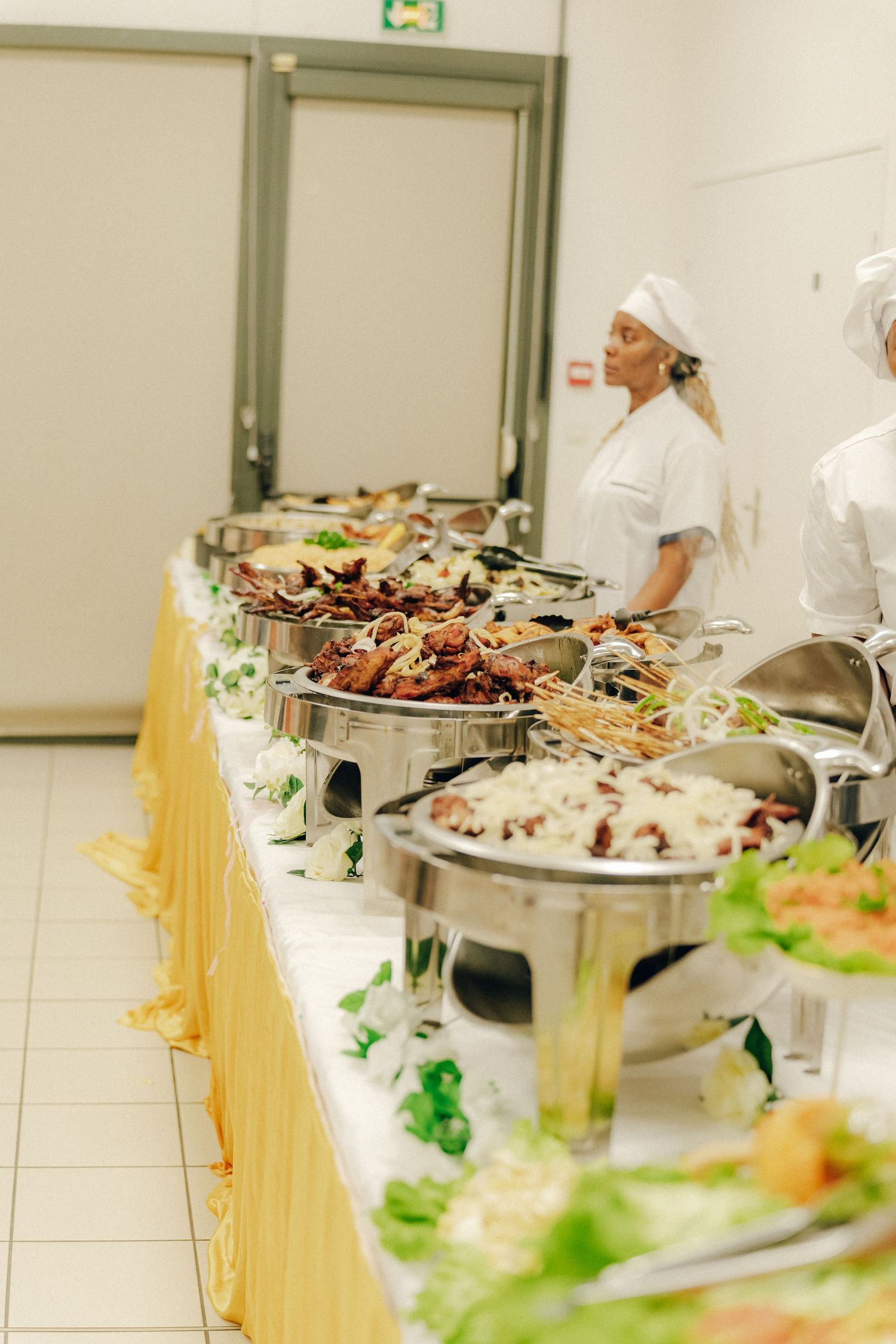 A buffet table with chafing dishes filled with catered food is attended by catering staff in white uniforms and chef hats.