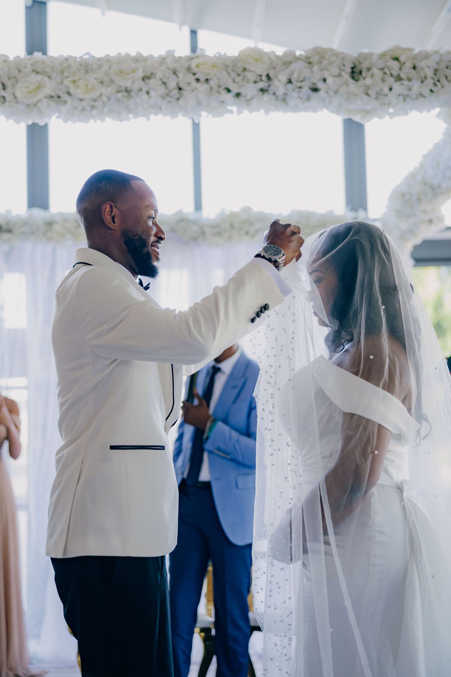 A touching moment during a wedding ceremony as the groom lifts the bride's veil under floral decorations.