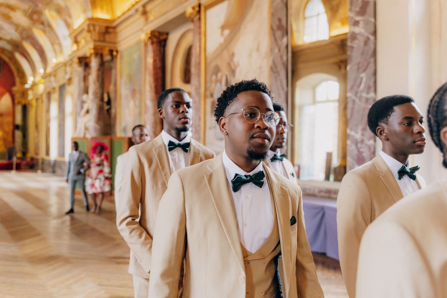 A group in formal attire poses in an elegant hallway with ornate architecture and arched windows.