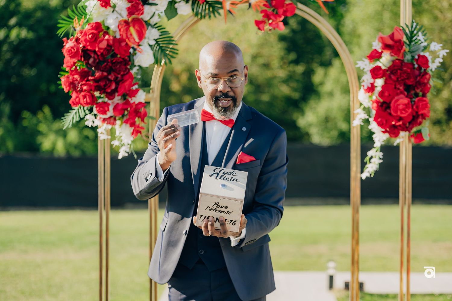 Wedding speaker at outdoor ceremony podium with red floral arch decorations.