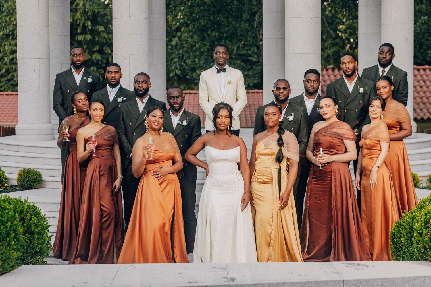 Wedding party in orange dresses and gray suits poses together on steps with columns.