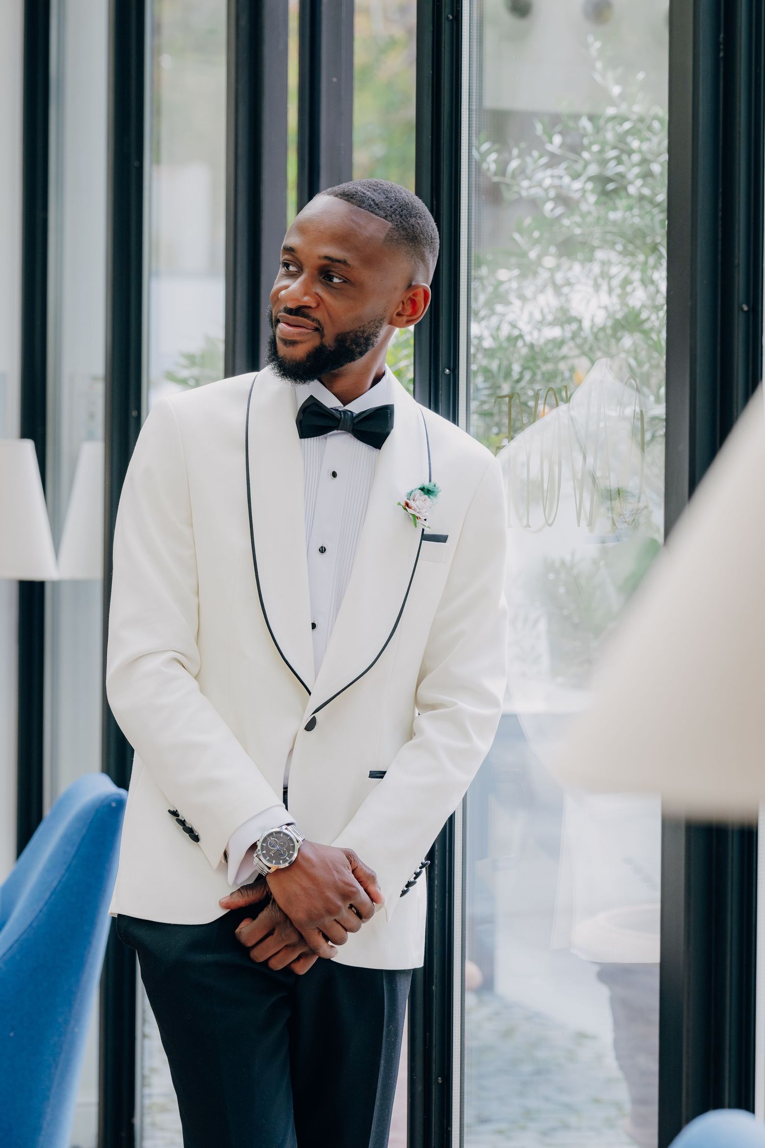 Two men in white tuxedo jackets and bow ties share a candid moment while standing by glass windows.