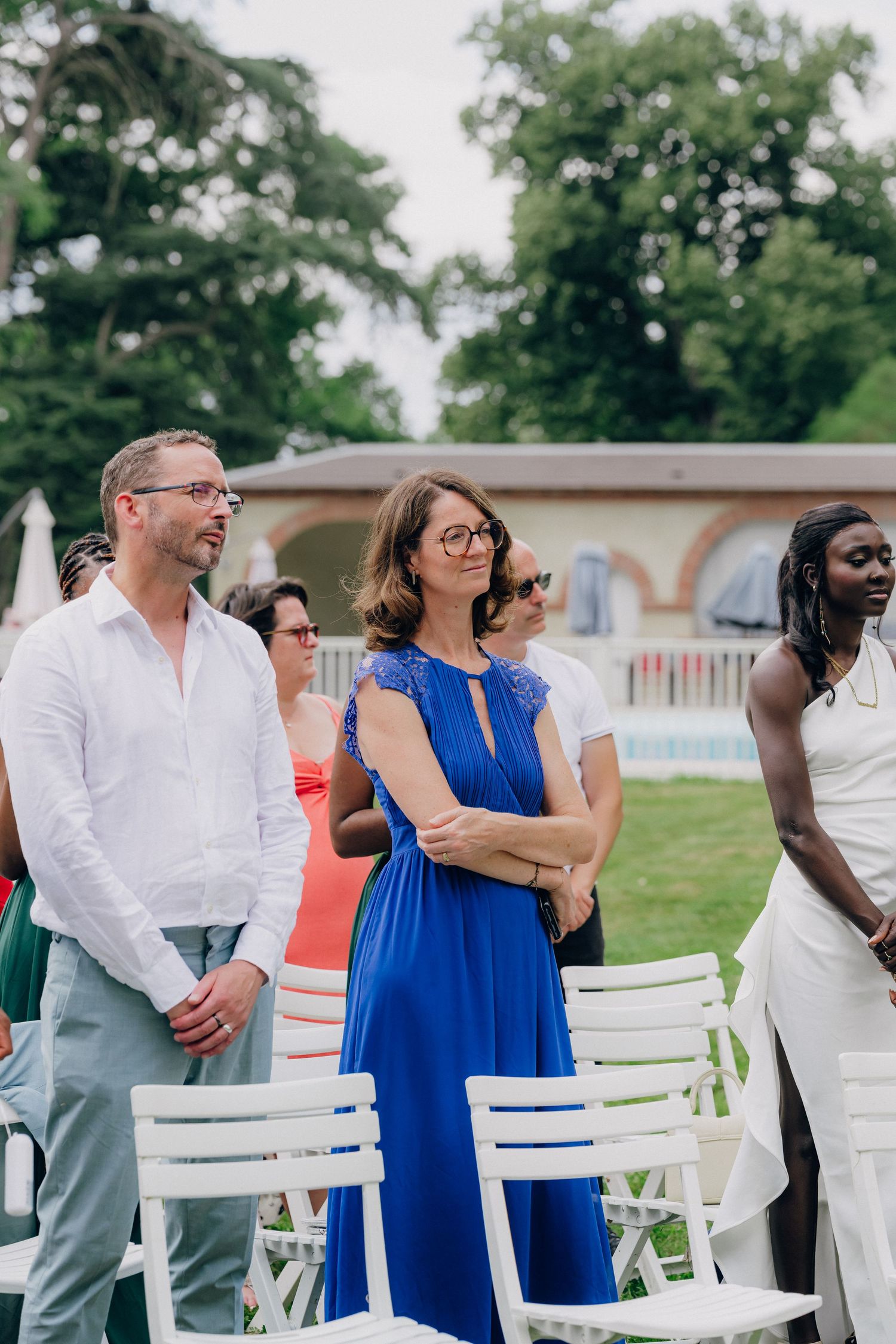 Wedding guests in colorful attire stand near white chairs during an outdoor ceremony.
