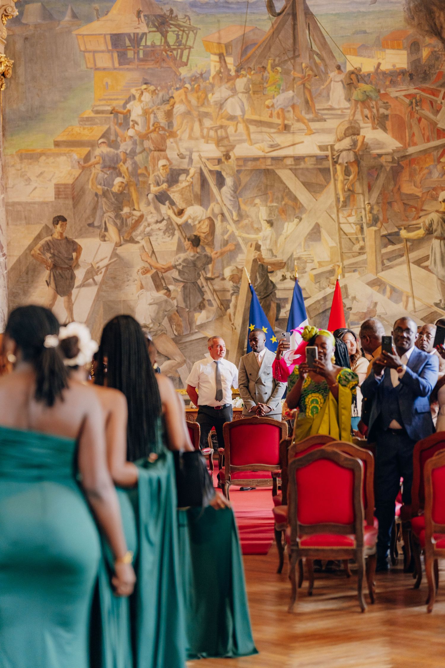 Formal wedding ceremony in an ornate historic hall with murals on the walls and guests in elegant attire.