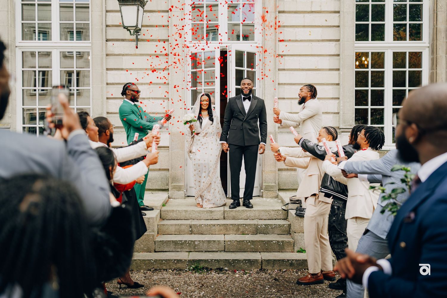 Wedding couple stands on steps while guests throw confetti in celebration outside historic building.