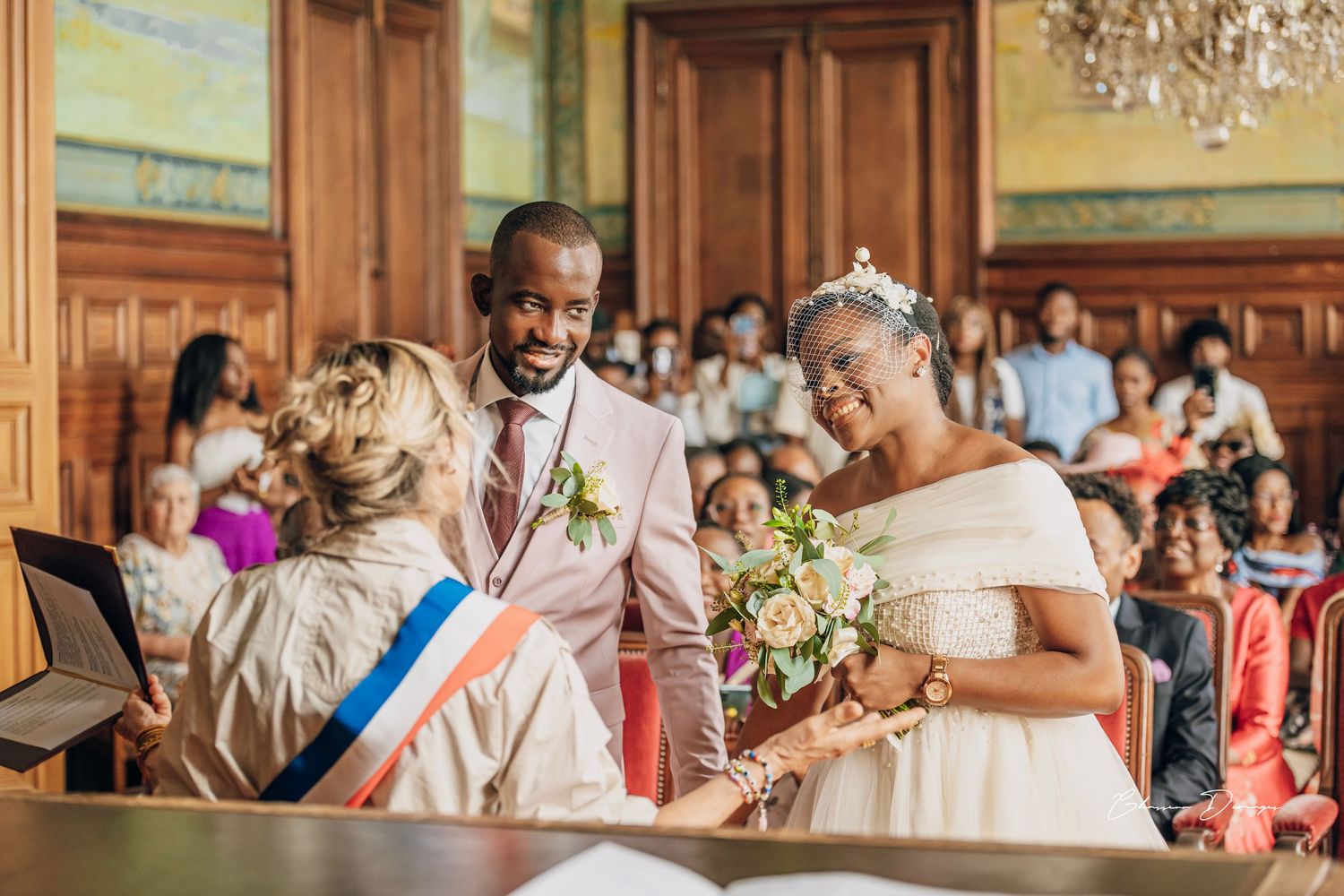 Civil ceremony taking place in wood-paneled room with ornate chandelier and flowers.