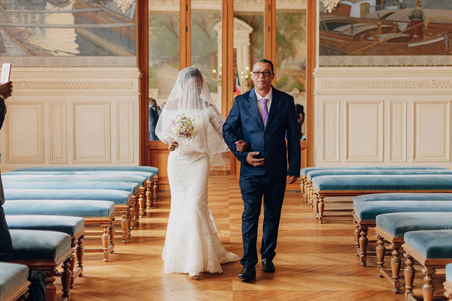 Newlyweds walking down aisle in historic venue with blue pews and decorative wall panels.