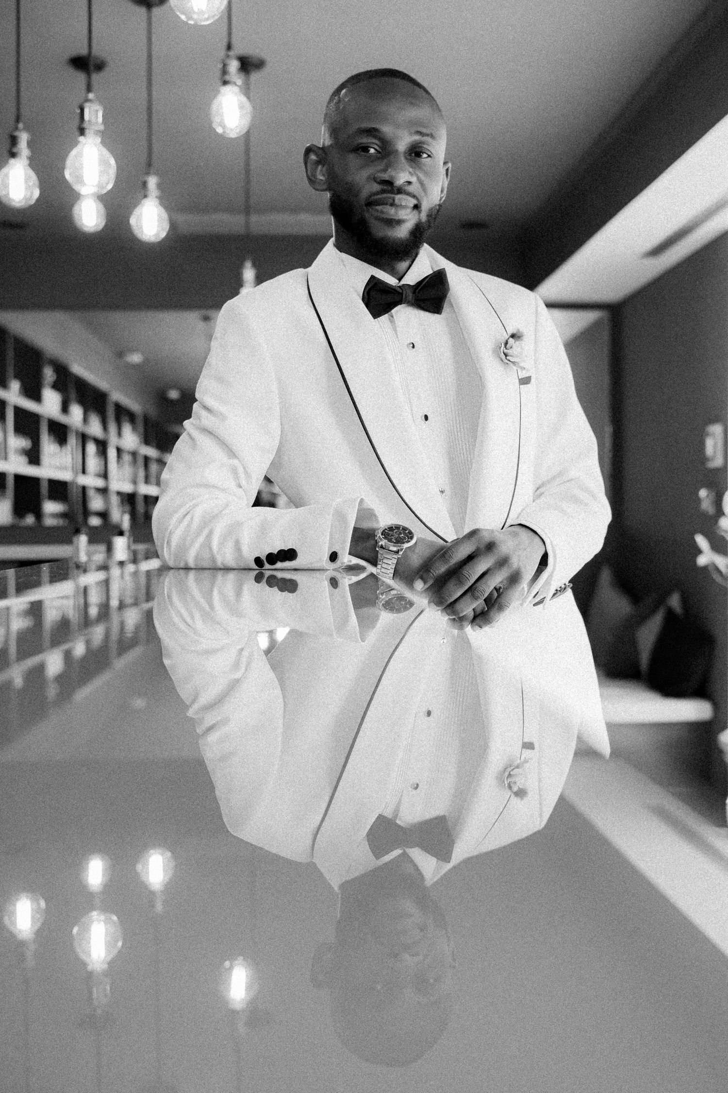 Person in white tuxedo jacket poses elegantly in modern hallway with hanging pendant lights.