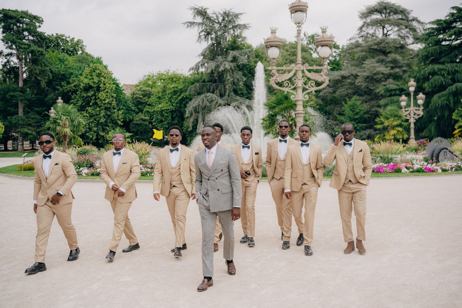 Group wearing matching beige outfits walks together through a park pathway on a cloudy day.