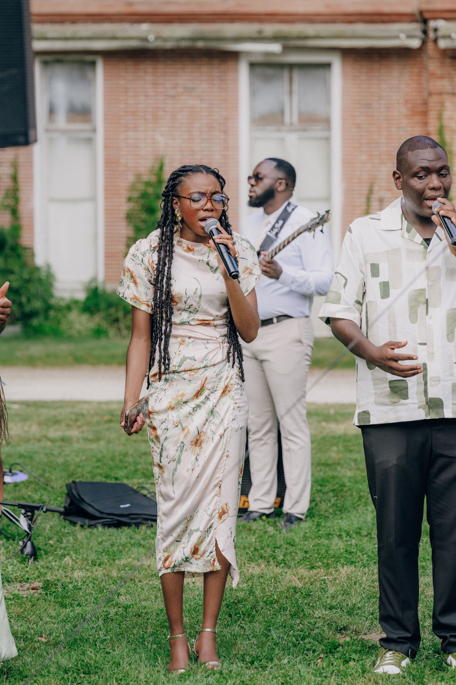 Wedding guests in formal attire mingle on a grassy lawn near a brick building.