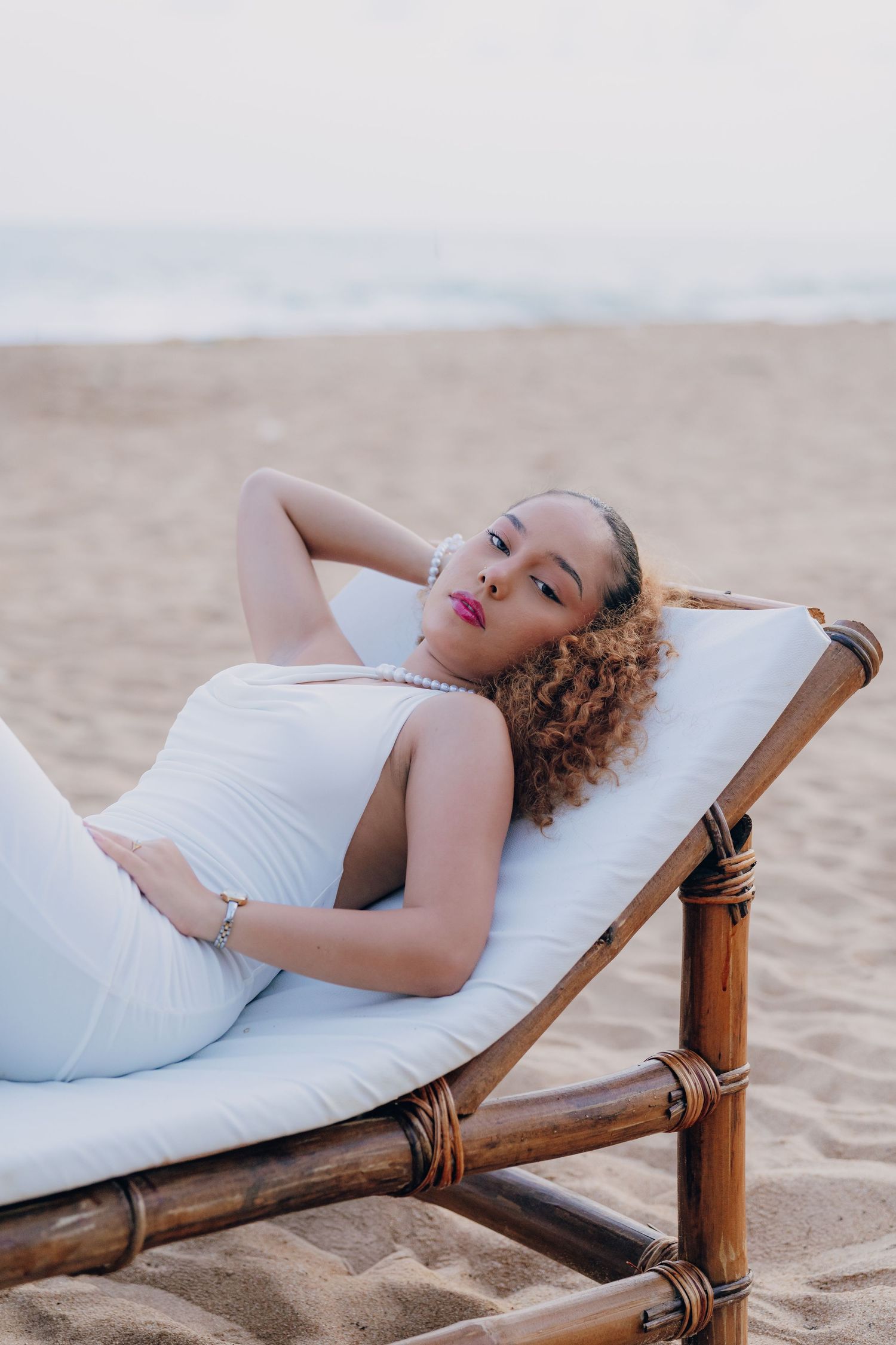 A young woman in a white dress reclines on a bamboo beach lounger on sandy shore, wearing pink lipstick and jewelry.